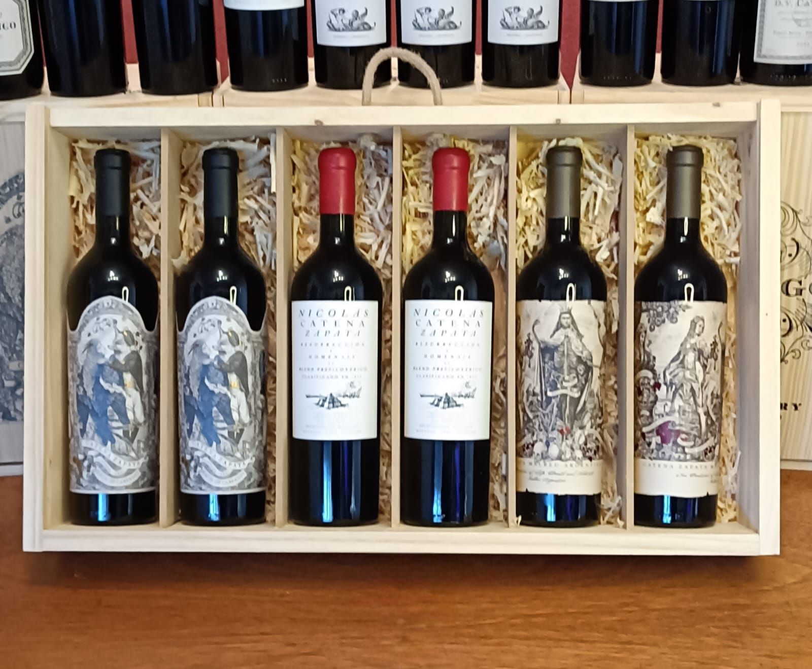 Seis botellas de vino tinto en una caja de regalo de madera sobre una mesa, con cuatro botellas más visibles encima.