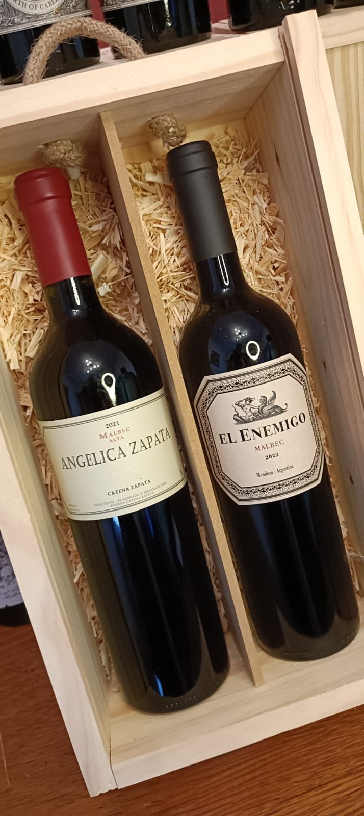 Dos botellas de vino tinto en una caja de regalo de madera, una con tapón rojo y otra con tapón negro.