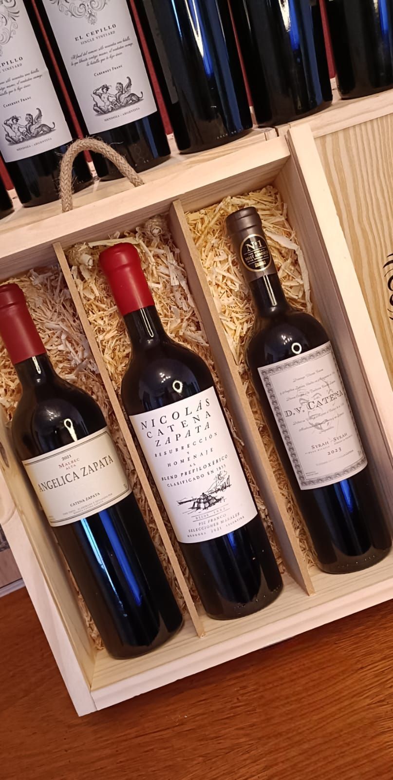 Caja de madera con tres botellas de vino tinto, dos etiquetas visibles y tapones de papel de aluminio rojo, sobre una mesa.