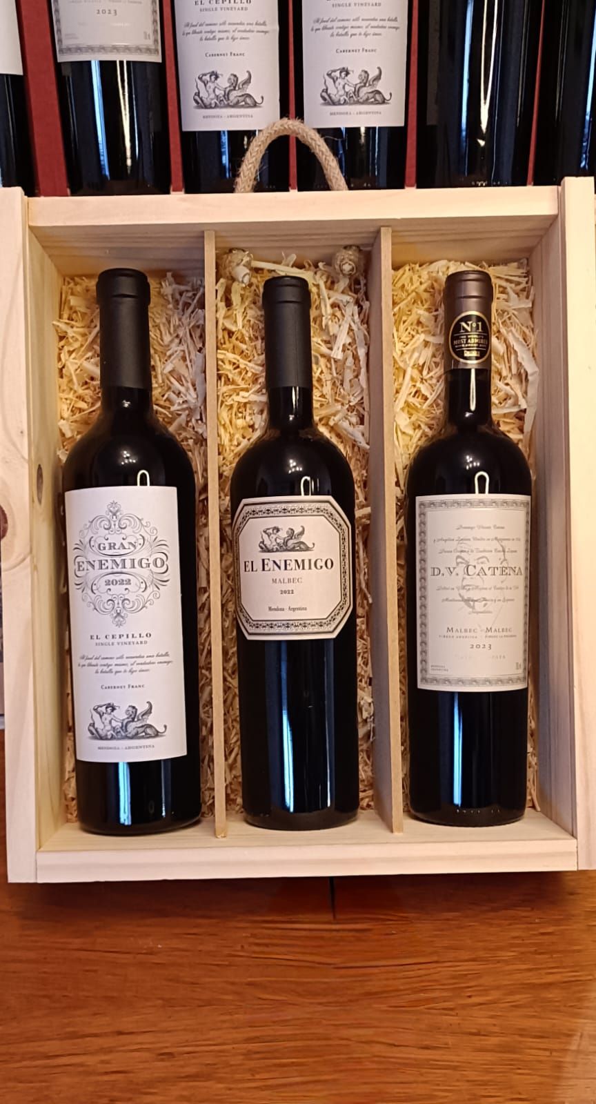 Tres botellas de vino en una caja de regalo de madera sobre una mesa, con libros en un estante detrás de ellas.