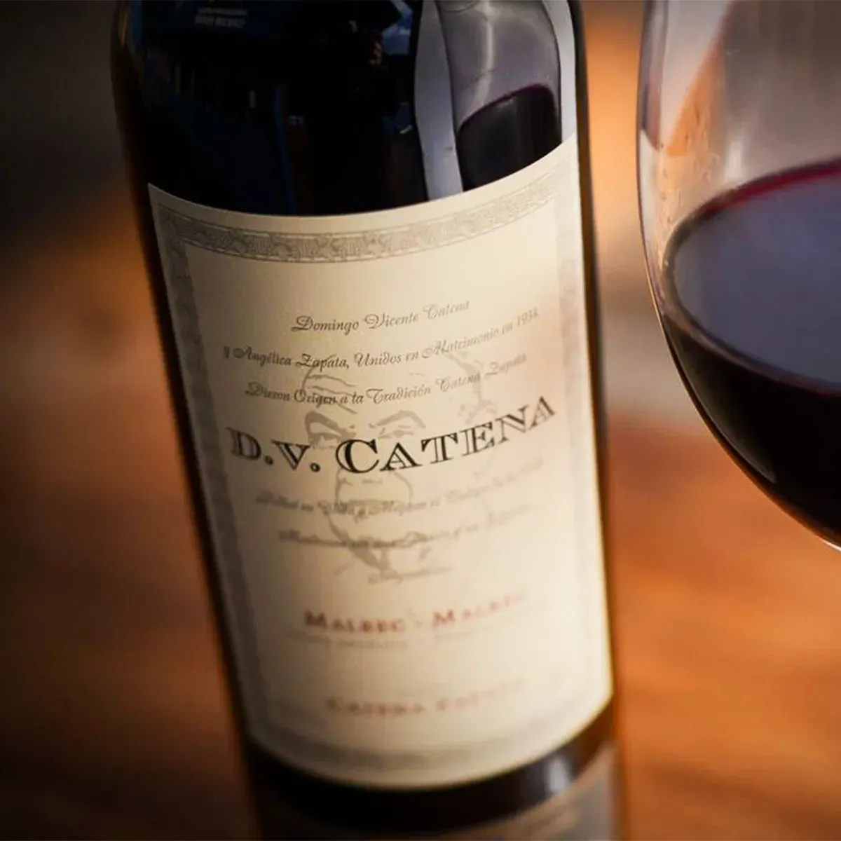 Una botella de vino tinto DV Catena junto a una copa sobre una mesa iluminada con luz cálida.