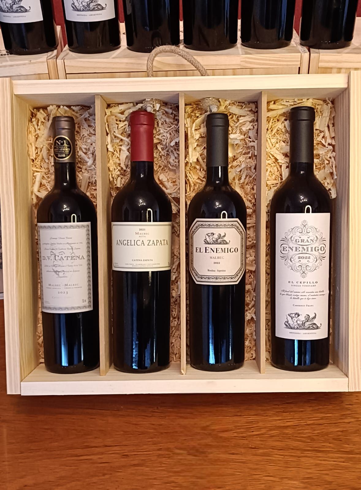 Cuatro botellas de vino en una caja de regalo de madera sobre una mesa, con más botellas en un estante detrás.