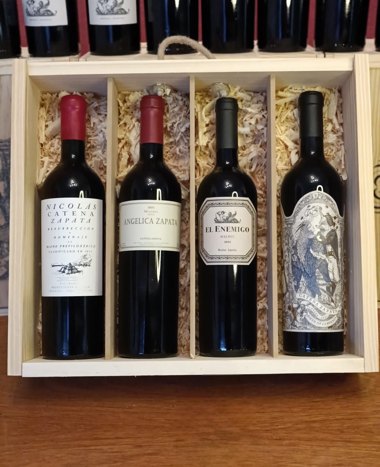 Cuatro botellas de vino tinto en una caja de regalo de madera con embalaje de paja y etiquetas decorativas.