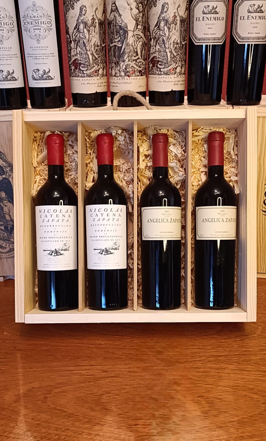Caja de madera para vino con cuatro botellas de vino tinto en la parte delantera y botellas adicionales en un estante detrás.