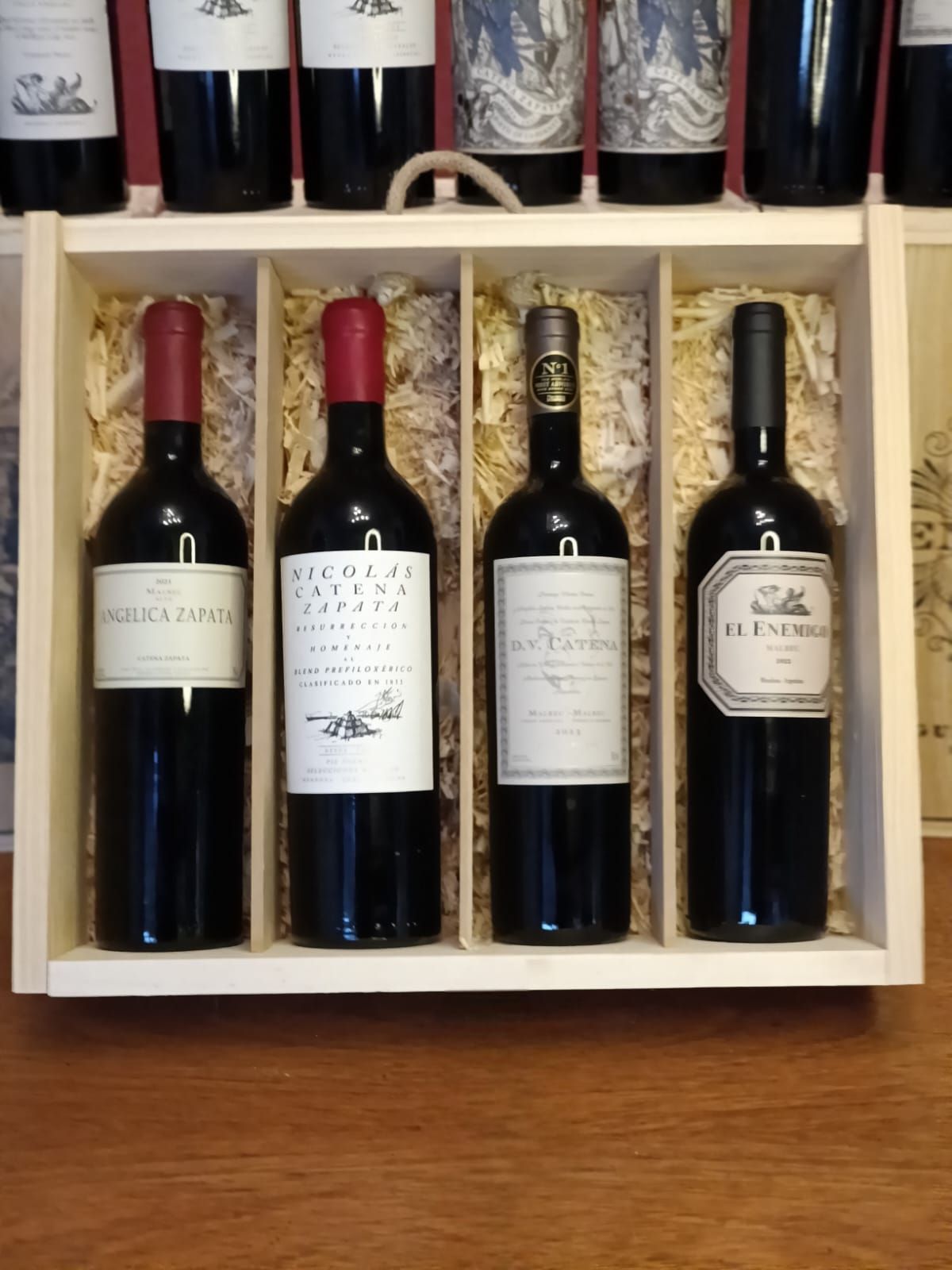 Sobre una mesa de madera, se puede ver una caja de madera abierta con cuatro botellas de vino tinto y sus etiquetas.