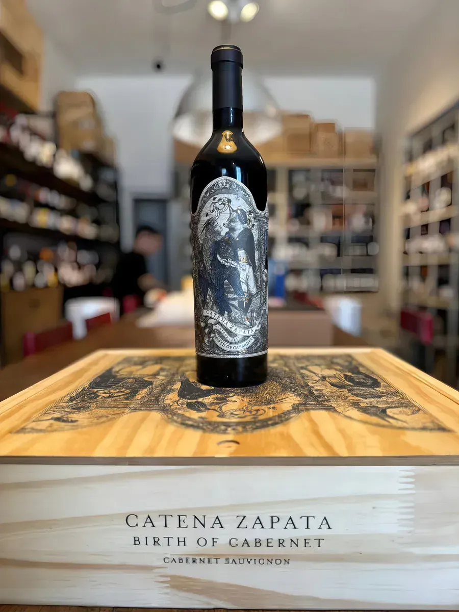 Tres botellas de vino oscuro con etiquetas ornamentadas dispuestas sobre una mesa negra sobre un fondo gris.