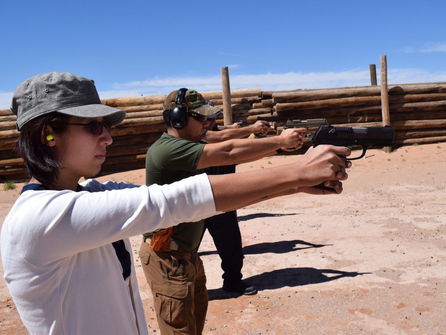 People at a shooting range aiming handguns. Bright day, red dirt, wooden barrier in background.