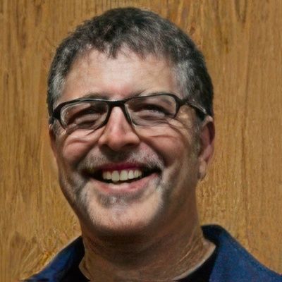 A man wearing glasses is smiling in front of a wooden wall.