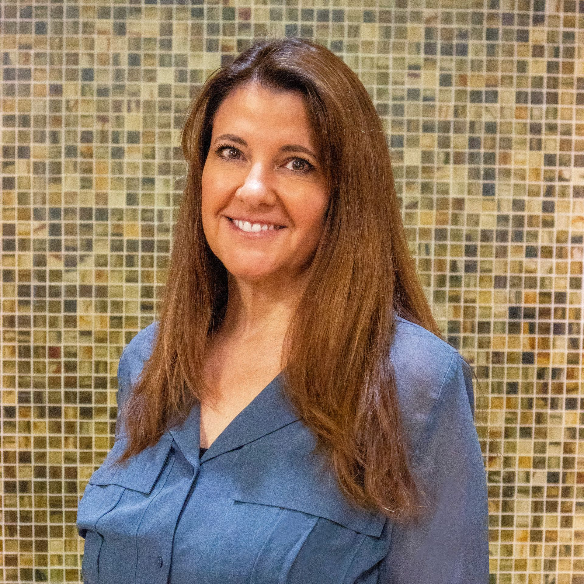 Woman with brown hair smiles, wearing a red long-sleeved shirt, near a window in an office setting.