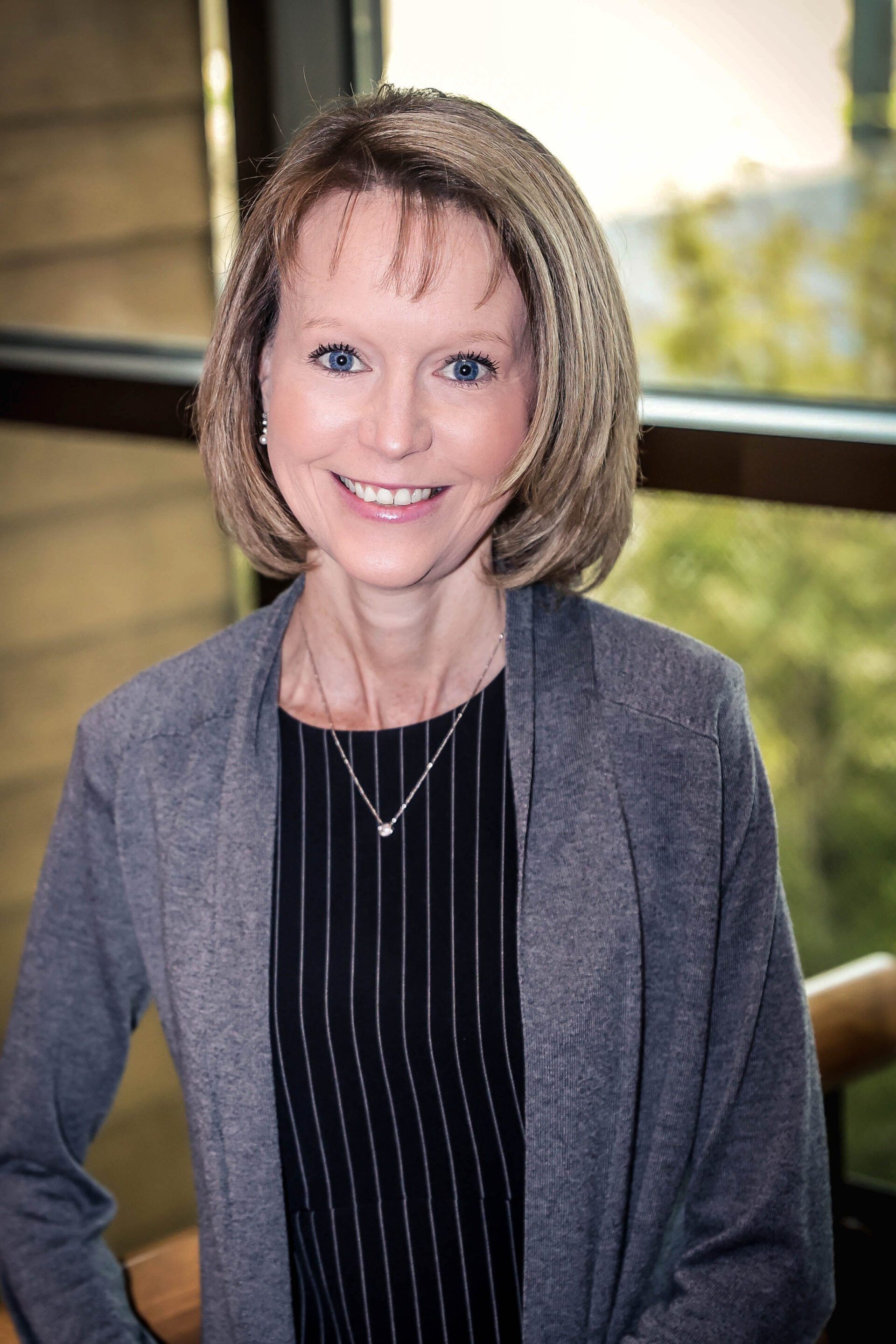 Woman in gray cardigan smiling, standing by a window, wearing a black and white striped top and a necklace.