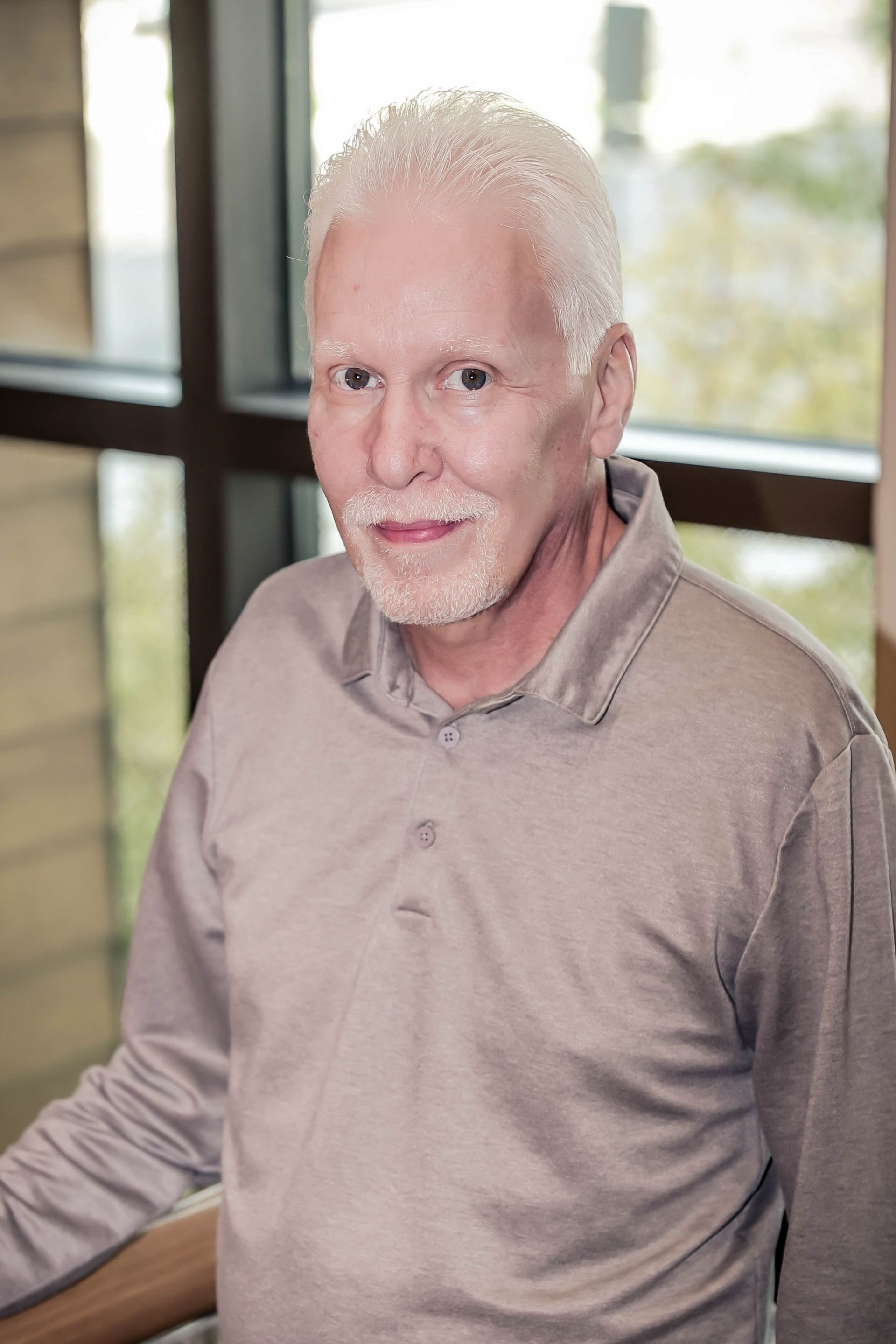Man with white hair and short beard wearing a grey polo shirt, smiling, standing indoors near a window.