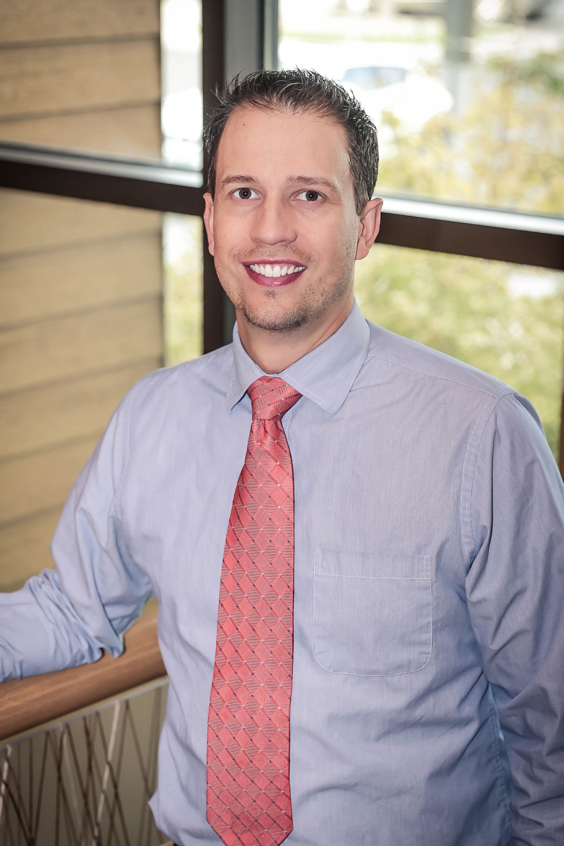 Man in blue striped shirt and red tie smiling indoors, leaning on a railing.