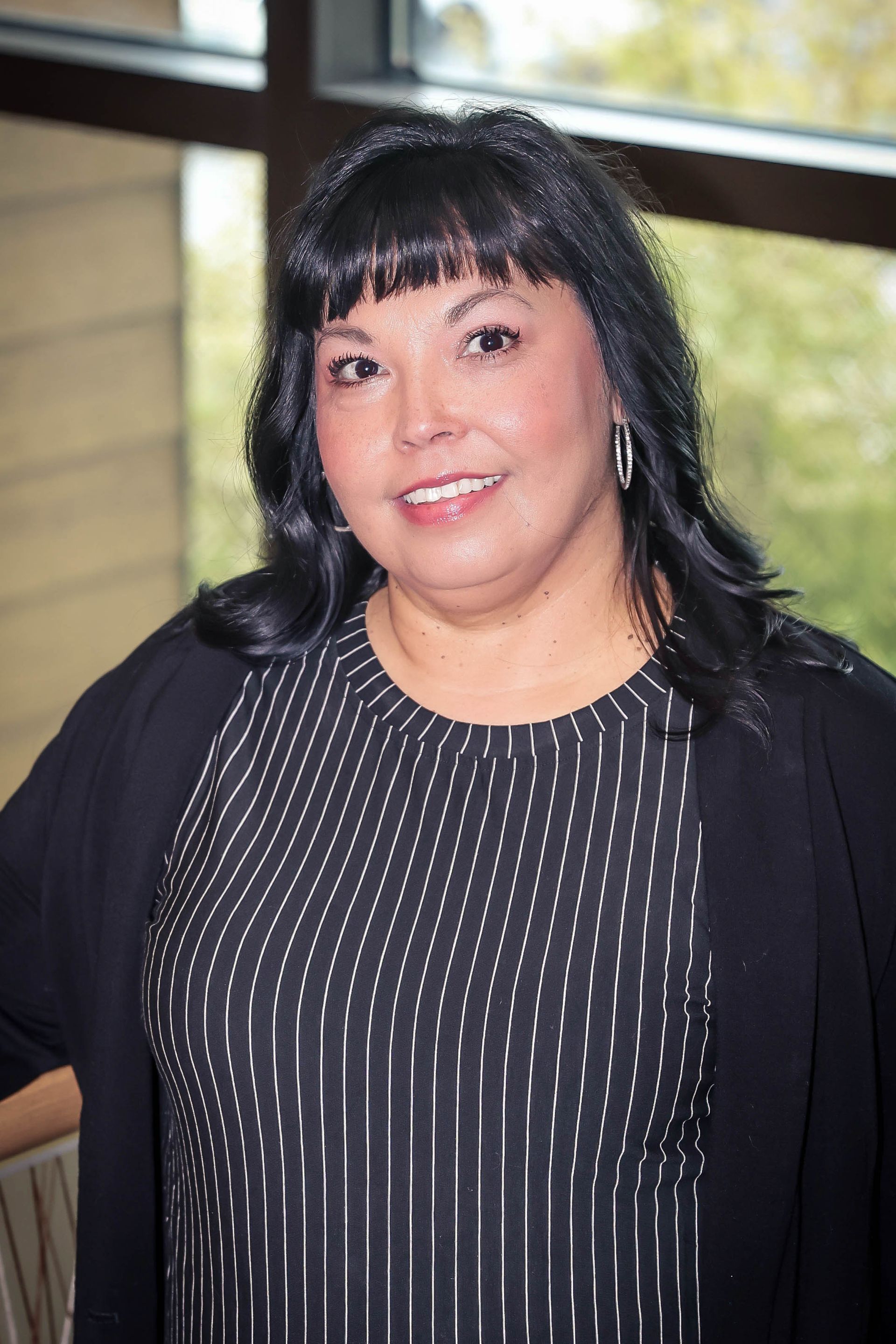 Woman smiling, black hair, wearing black top, black jacket, and hoop earrings, standing indoors.