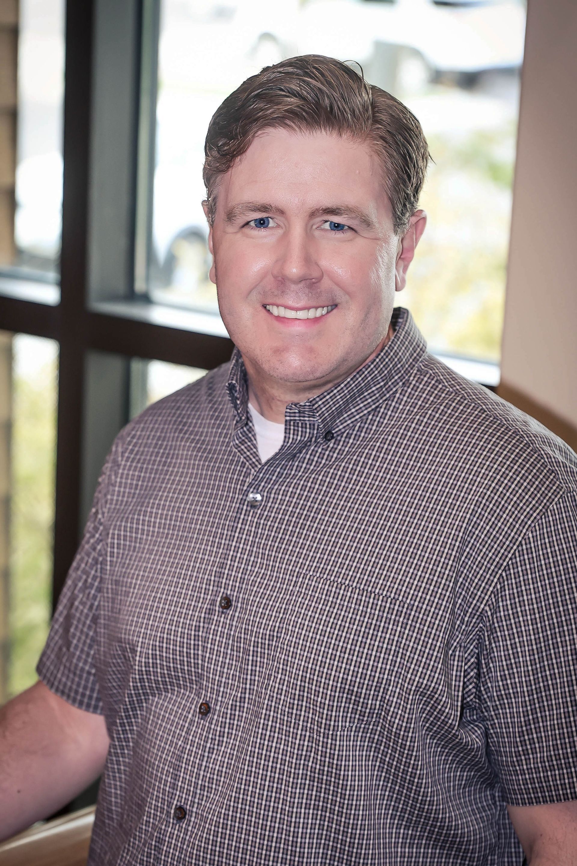 Man smiling, standing by window, wearing a button-down shirt.