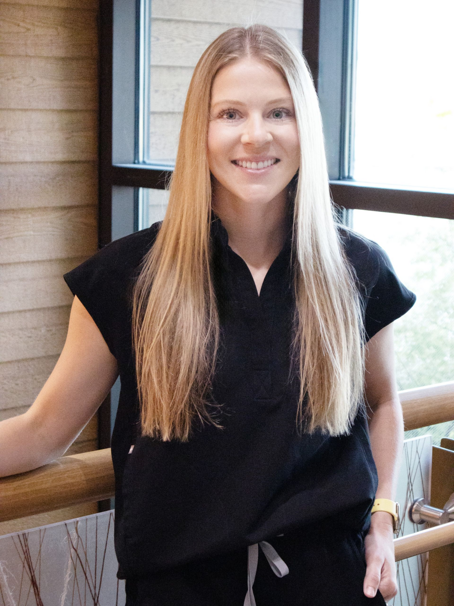 Woman with long blonde hair, smiling, wearing black scrubs, standing near a wooden railing and window.