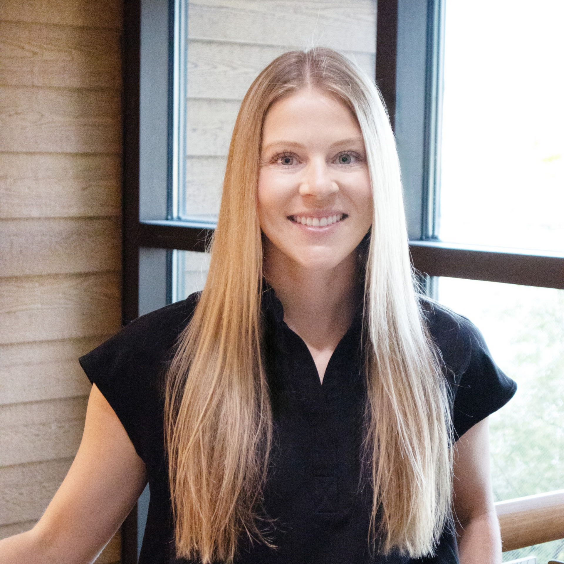 Woman with long blonde hair smiles, wearing a black top, by a window with wood siding.
