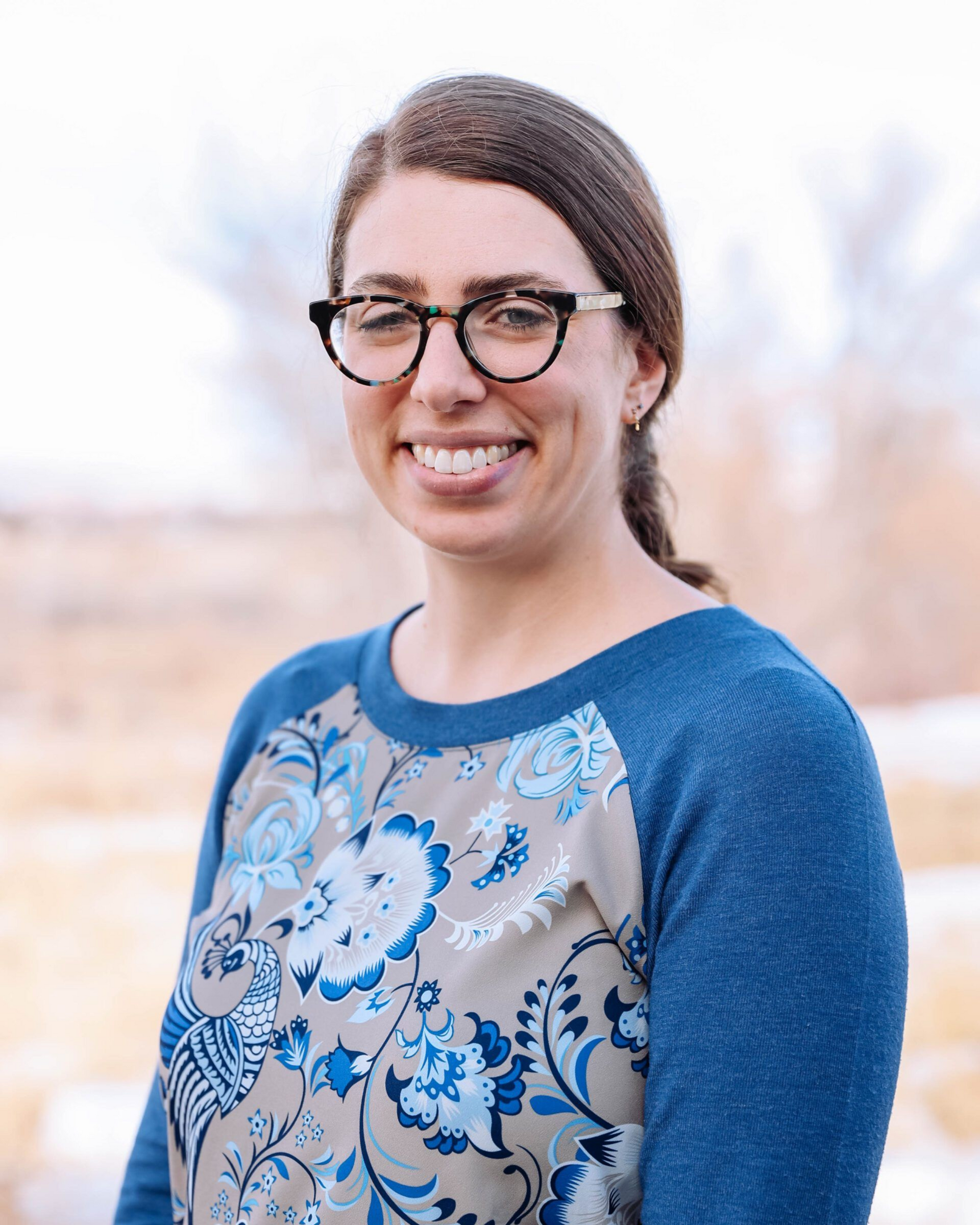 Woman with glasses smiles, wearing blue floral sweater, against a blurred outdoor background.