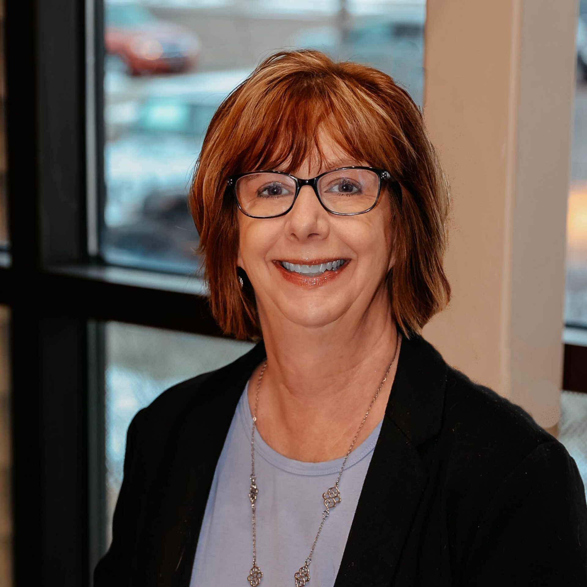 Woman with glasses smiling in front of a window, wearing a black blazer over a light blue shirt.