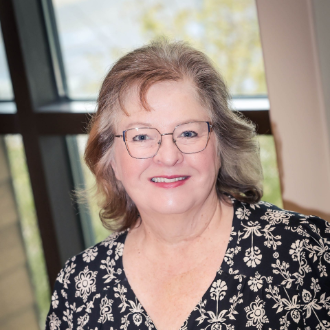 Woman with glasses smiles at the camera in a room with a window. She wears a floral blouse.