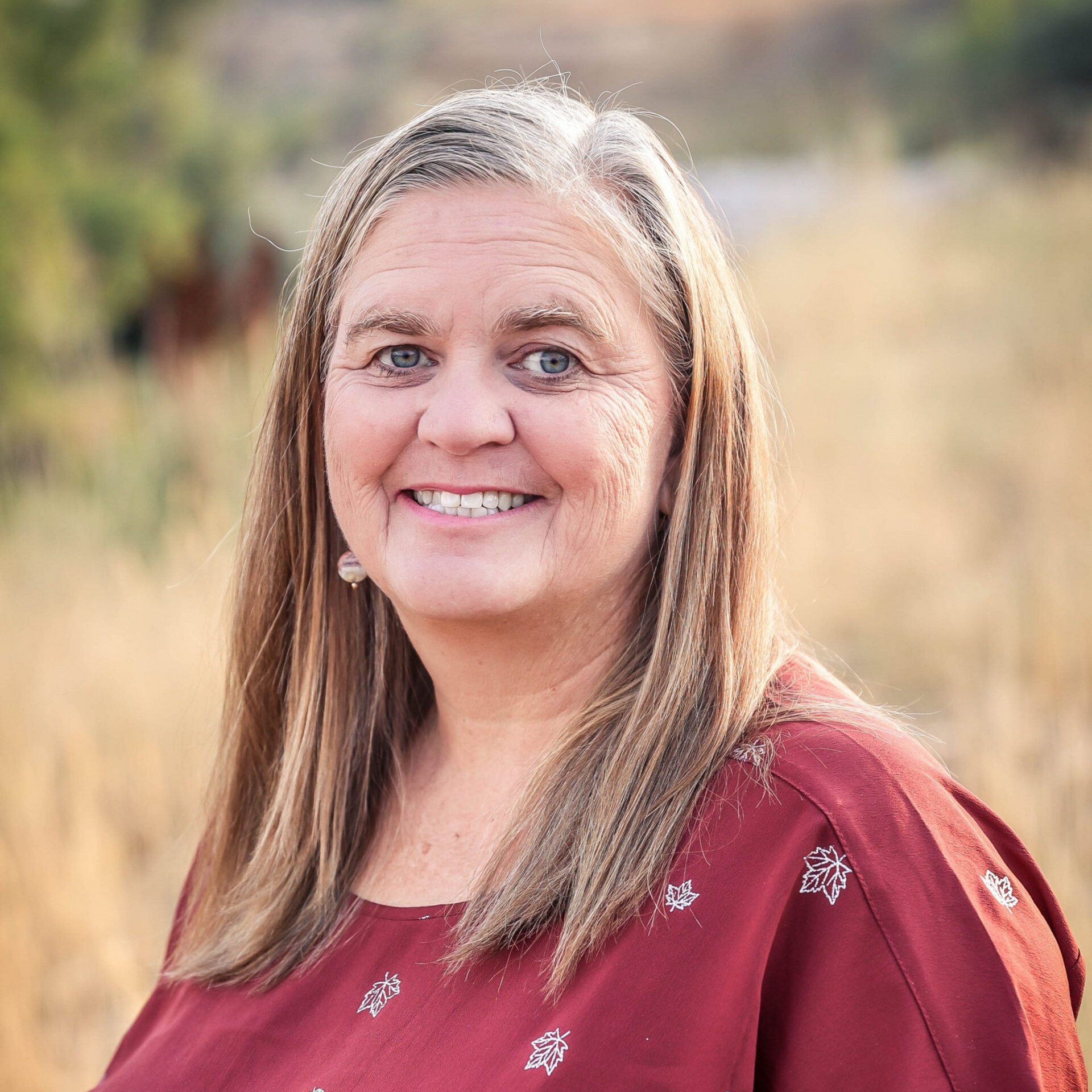 Woman with long gray-streaked hair smiles, wearing a burgundy top; outdoors with dry grass in the background.