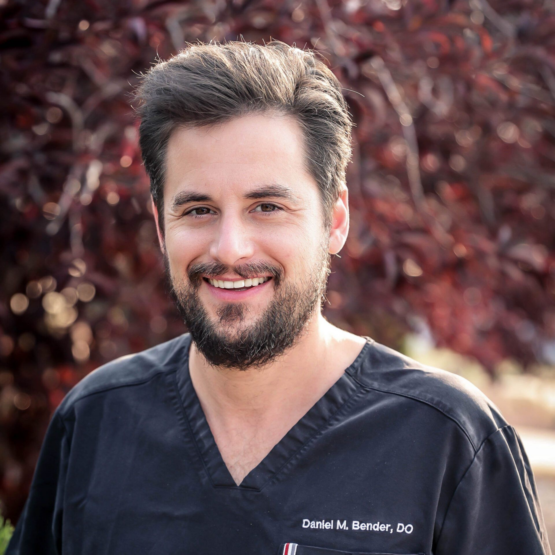 Man in black scrubs smiles, standing in front of red-leafed tree. Name tag reads