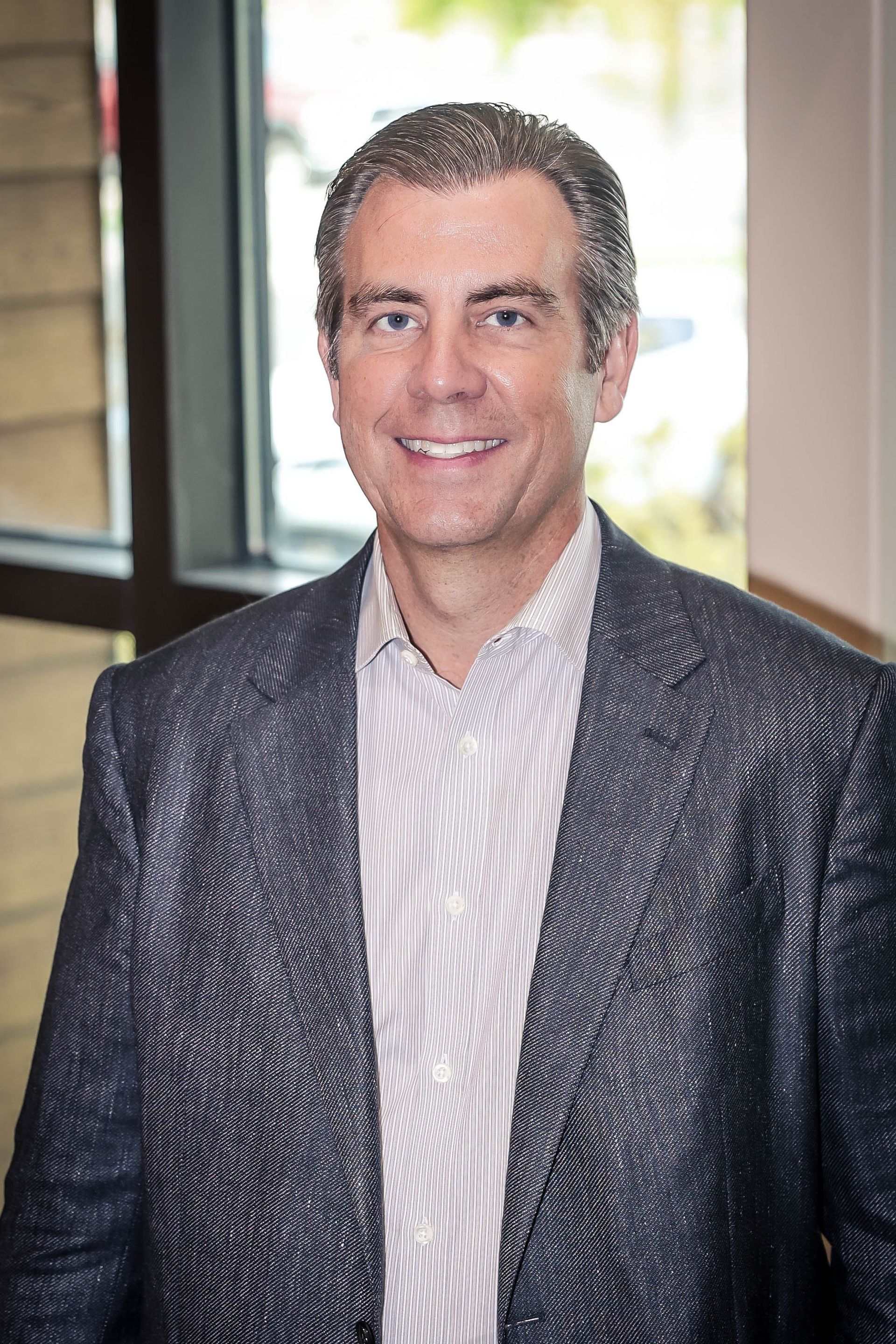 Man in blazer and striped shirt, smiling, by a window.