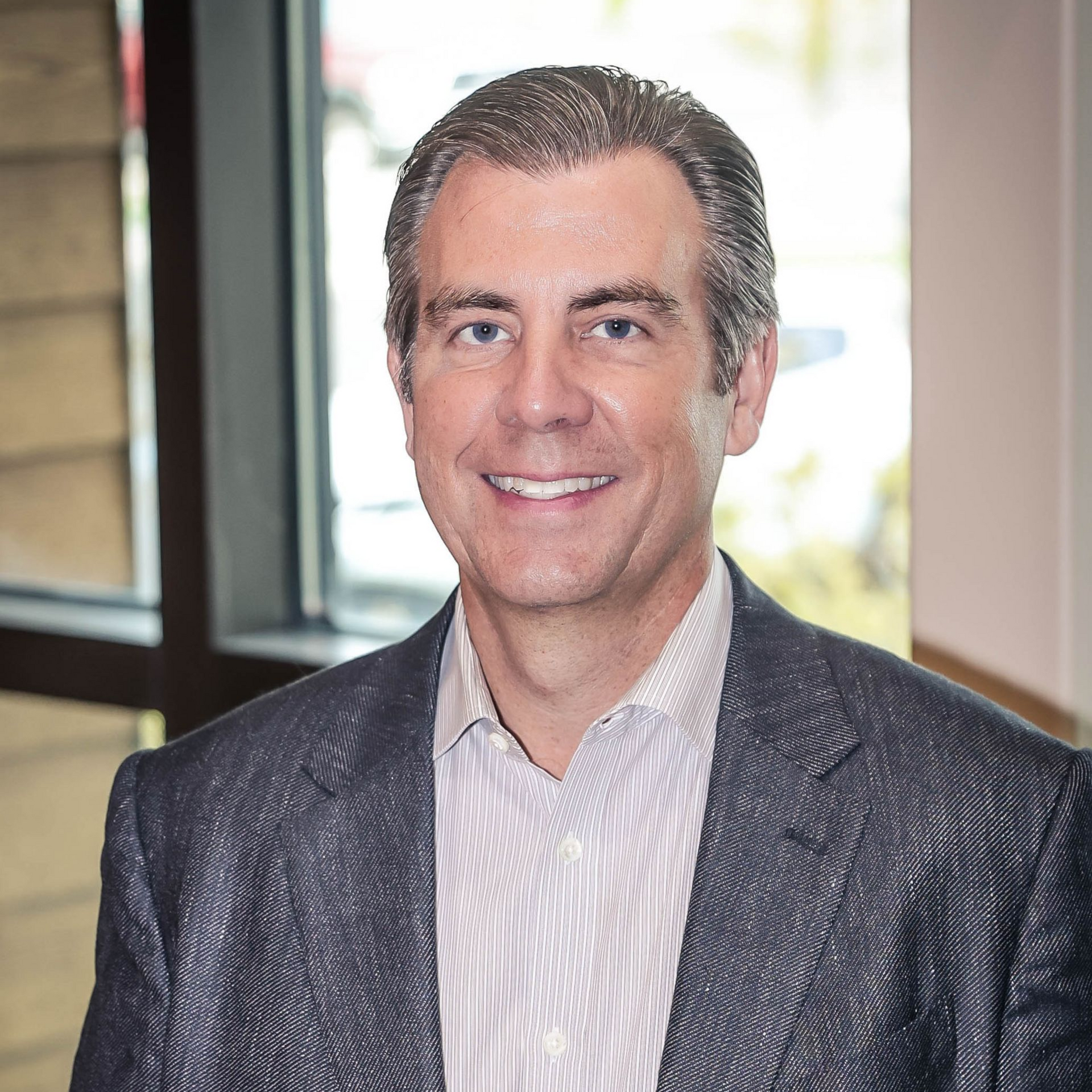 Man in a gray suit and striped shirt smiles indoors, near a window.
