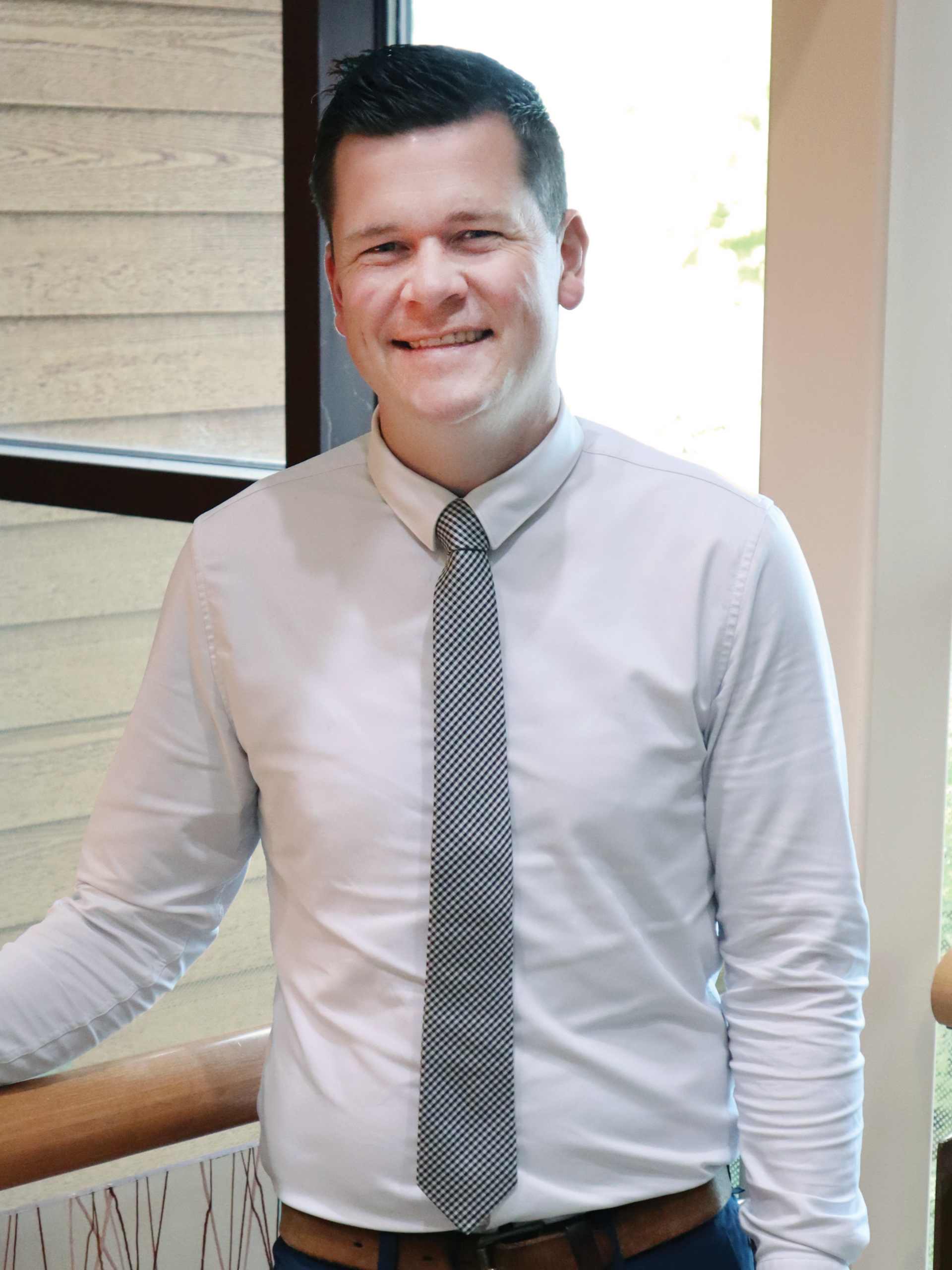 Man in a white shirt and tie smiles, standing by a wooden handrail near a window.