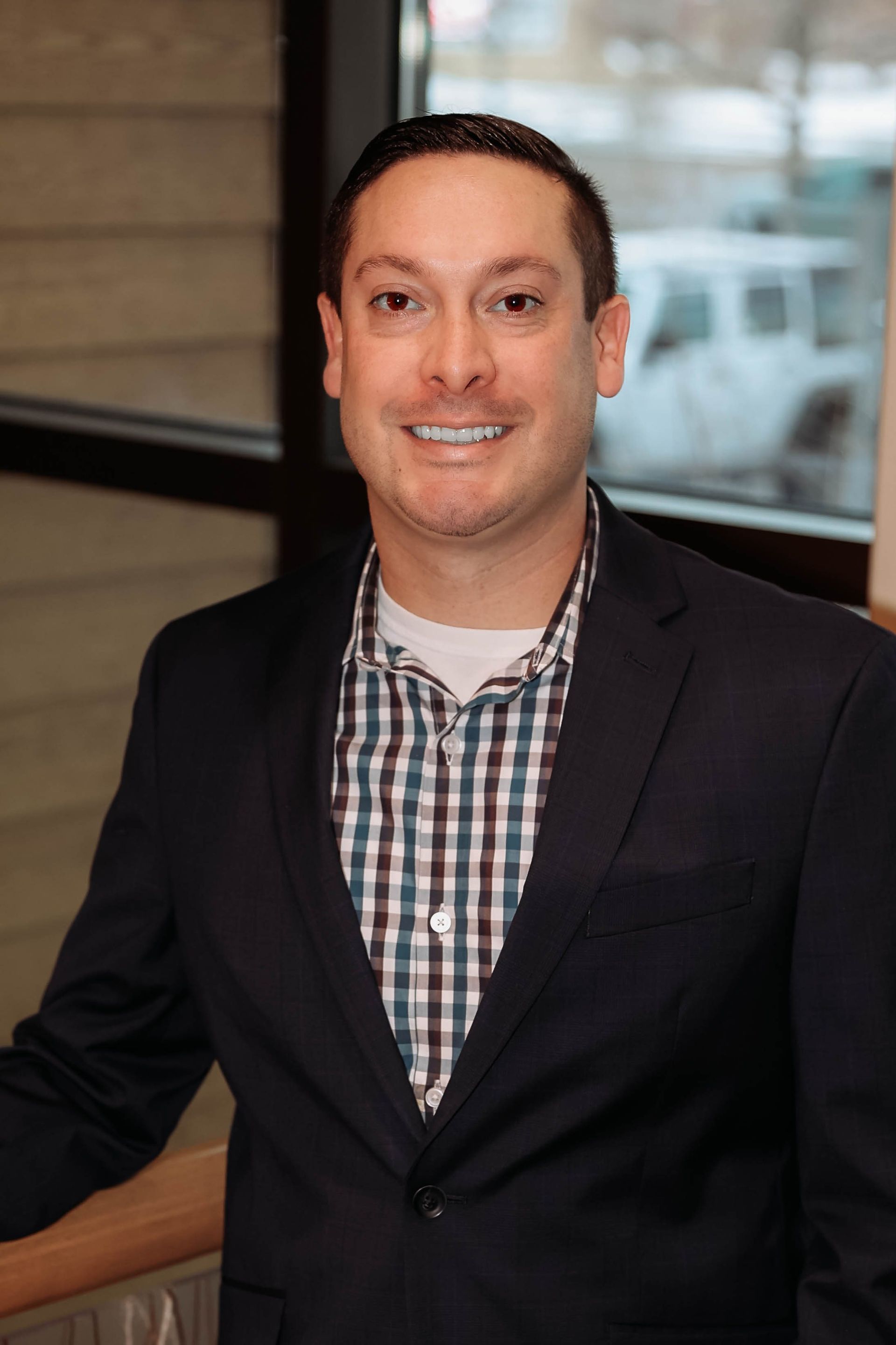 Man in a blazer and plaid shirt smiles indoors, leaning against a railing near a window.