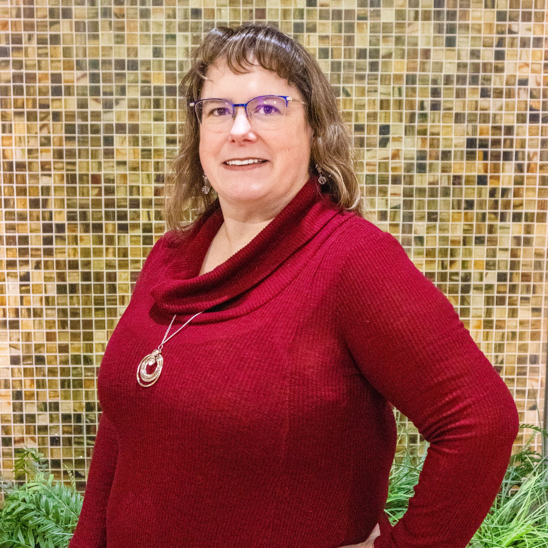 Woman wearing glasses, smiling, blue necklace, standing indoors, hands on railing.