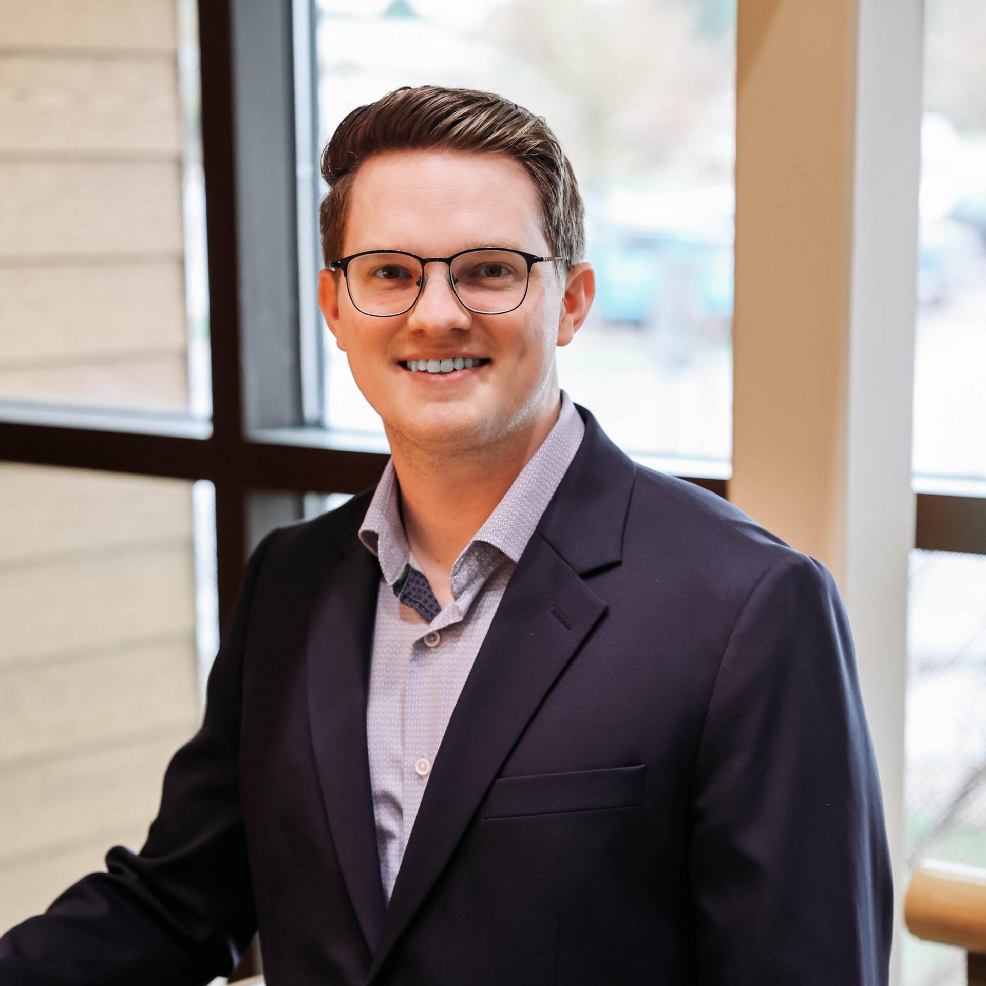 Man in glasses and navy blazer smiling near a window, natural light.