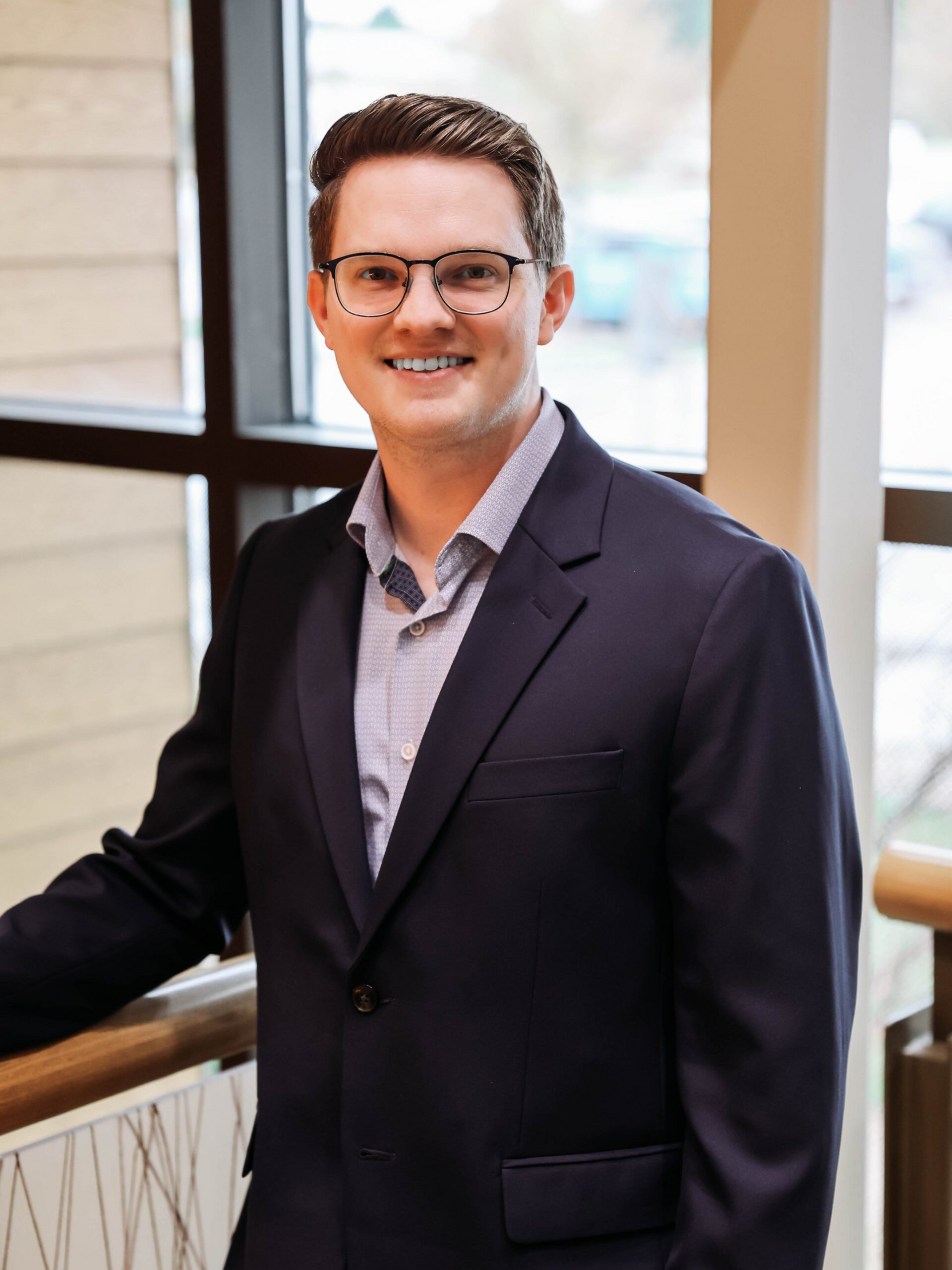 Man in a navy blazer and glasses smiles, leaning on a railing near a window.