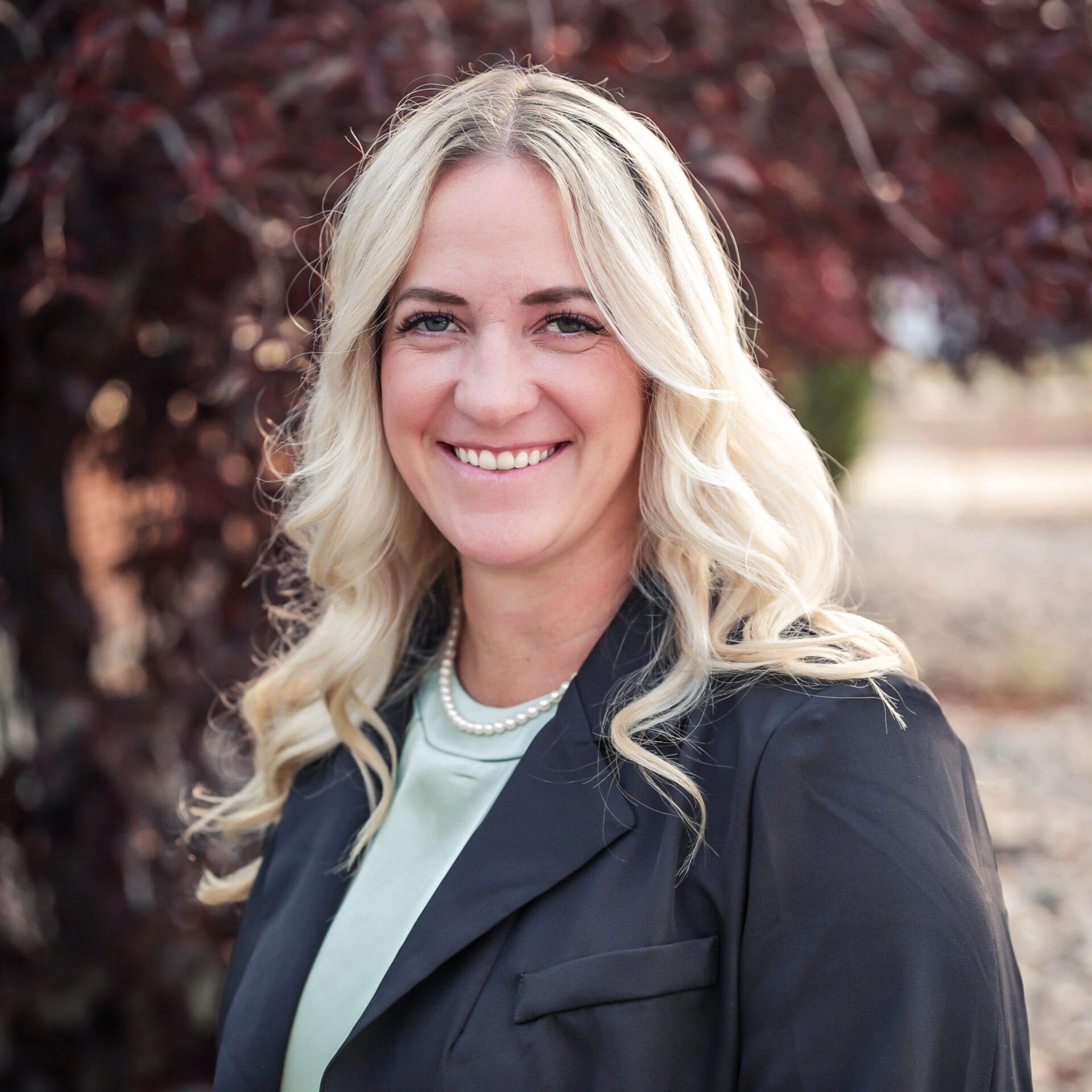 Blonde woman smiling, wearing a black blazer and pearl necklace, in front of a tree.