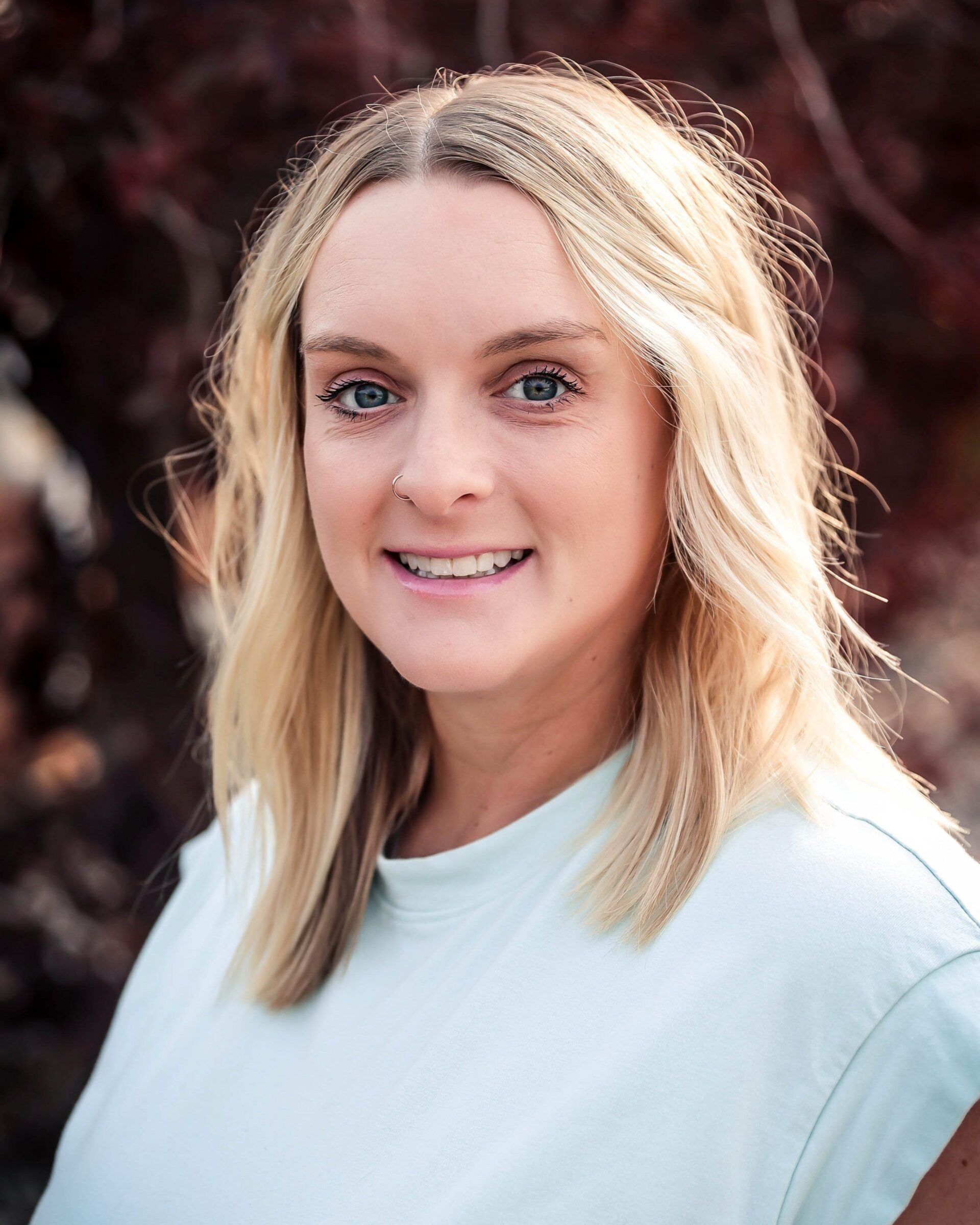 Blonde woman with blue eyes smiles, wearing a light blue shirt.