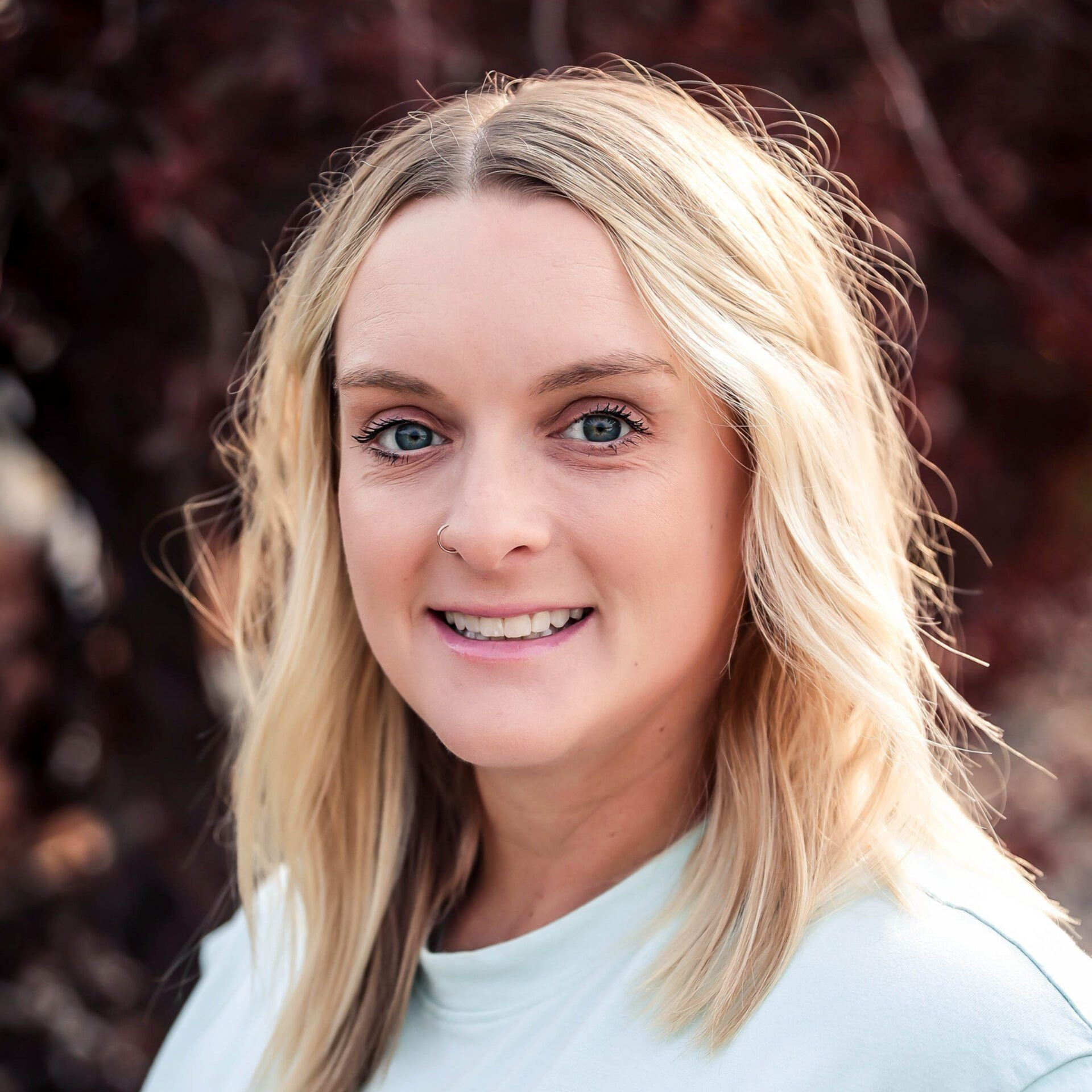 Blonde woman with blue eyes smiles, outdoors with blurred fall foliage background.