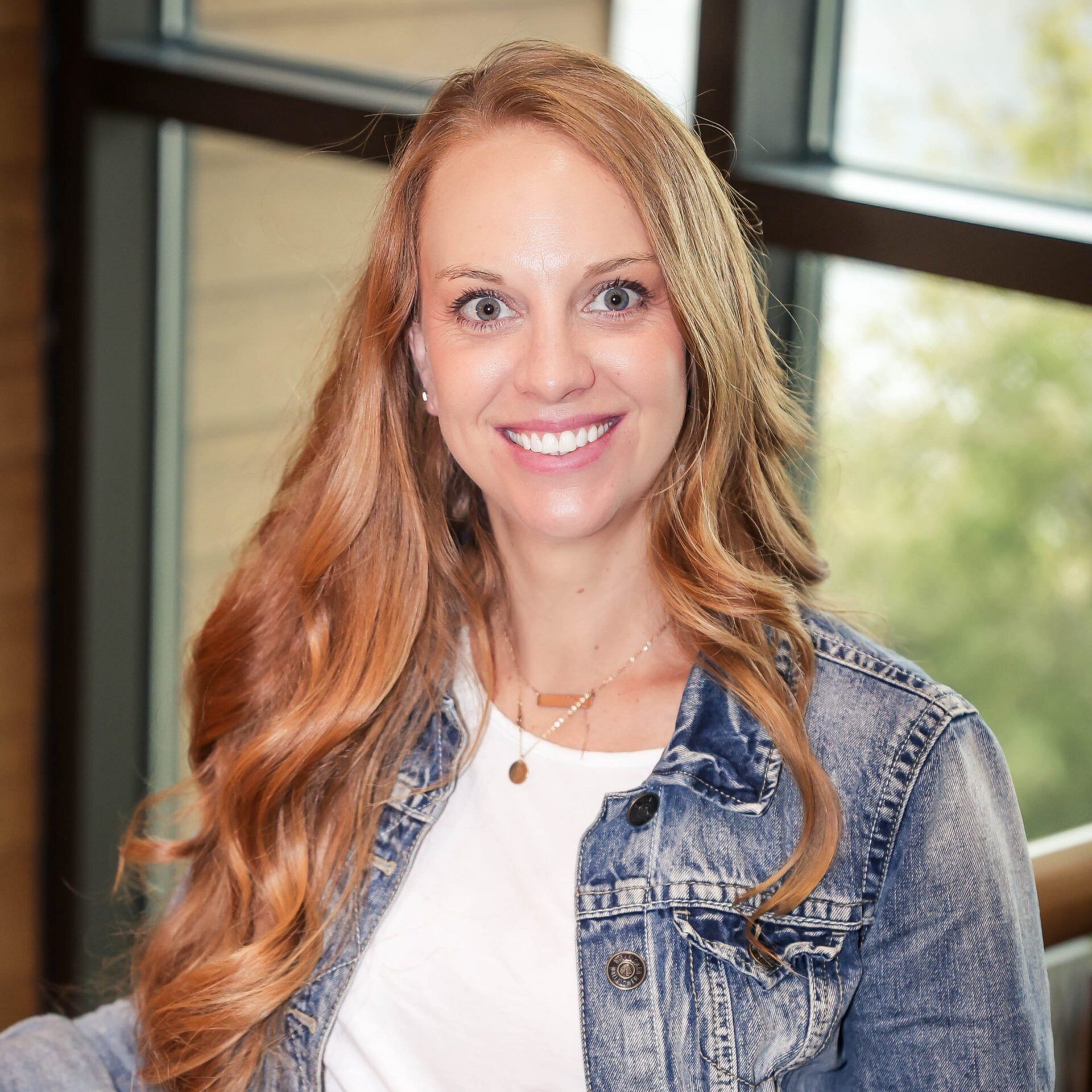 Woman with long auburn hair smiles, wearing a denim jacket and white shirt indoors near a window.