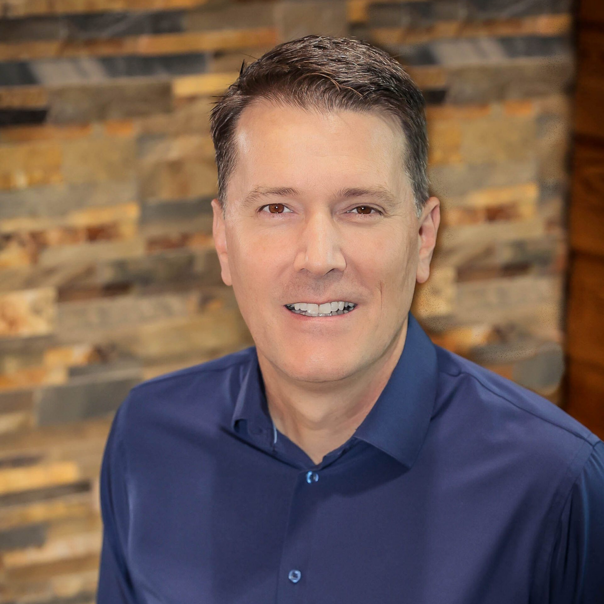 Man in blue shirt smiles in front of a stone wall.