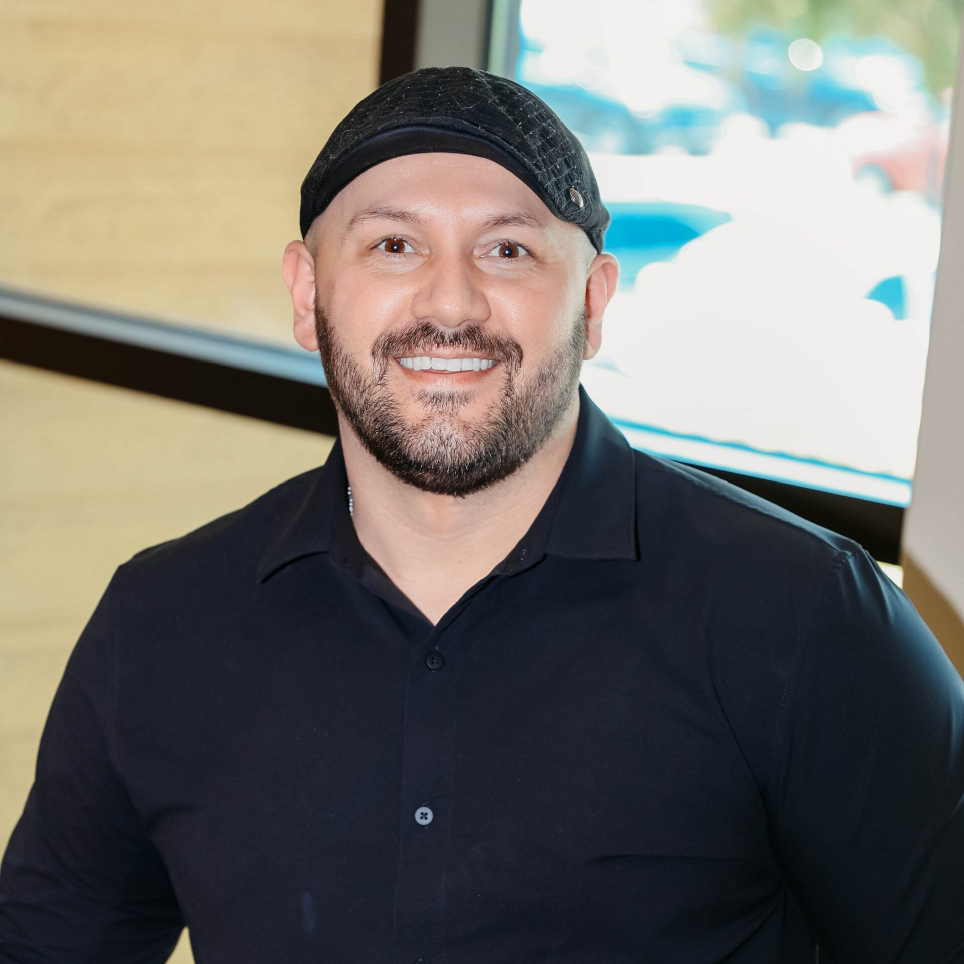 Man with a beard, wearing a black cap and shirt, smiling, in front of a window.