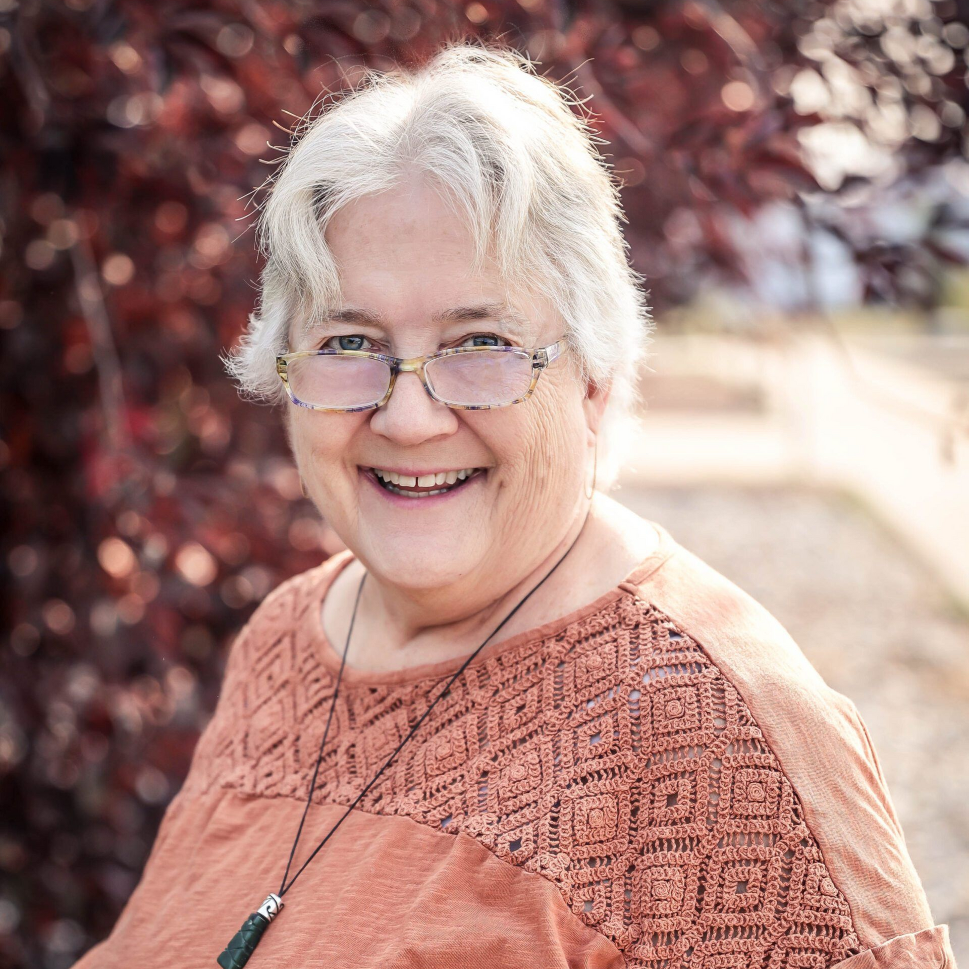 Woman smiling outdoors, wearing glasses and orange top; red and purple foliage in the background.