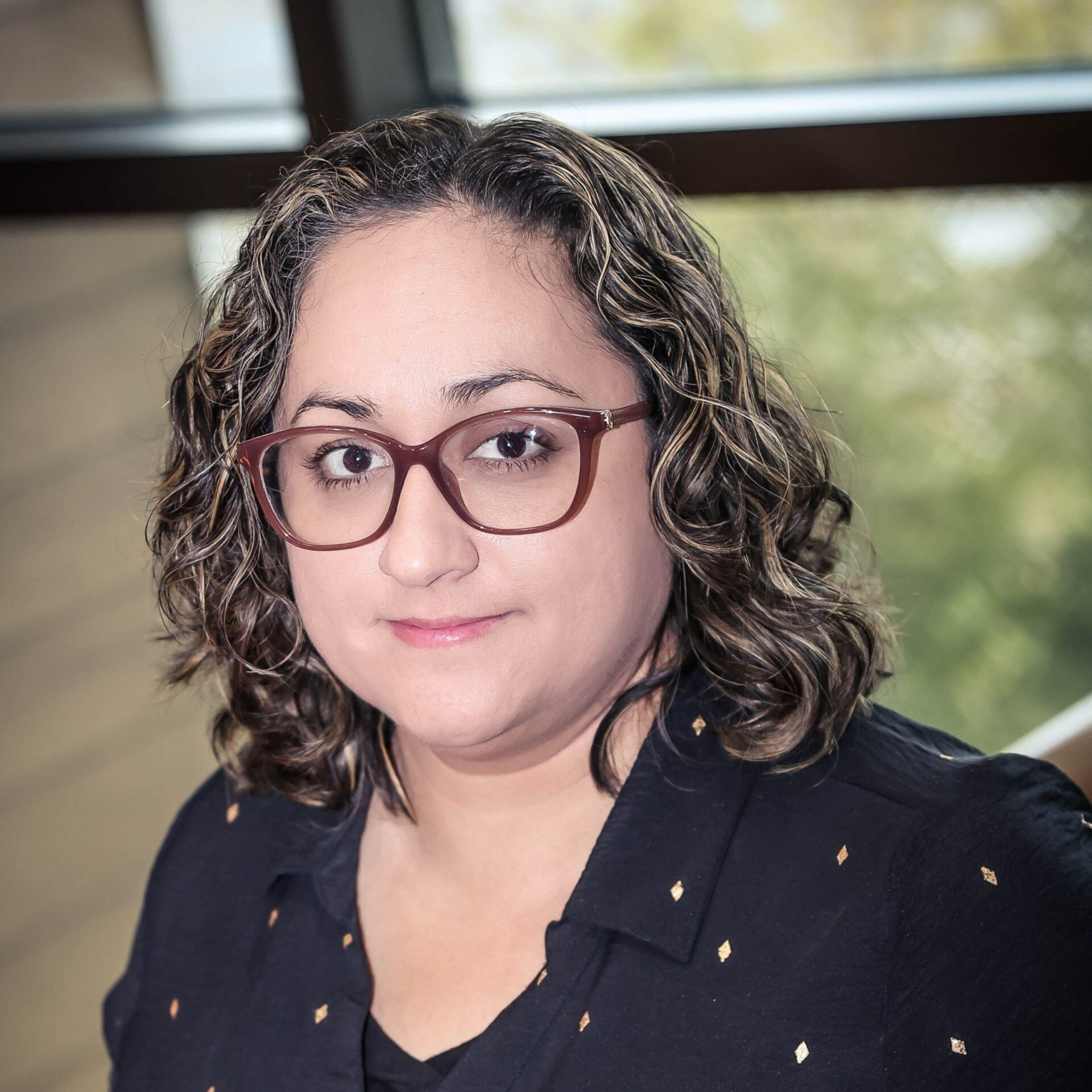 Woman with curly brown hair wearing glasses, black shirt with gold dots, looking at the camera.