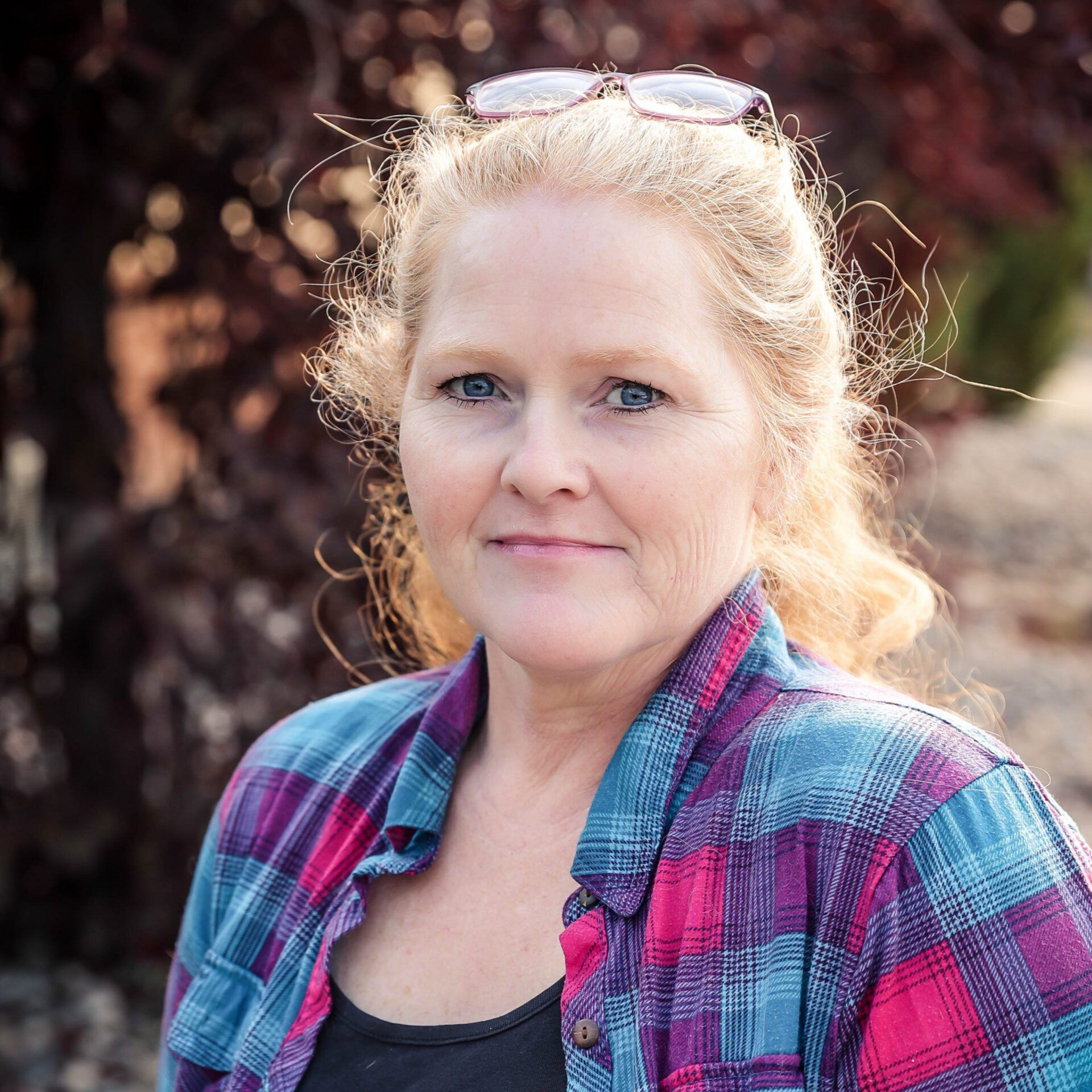 Woman with blonde hair wearing a blue and pink plaid shirt, smiling, with glasses on her head.