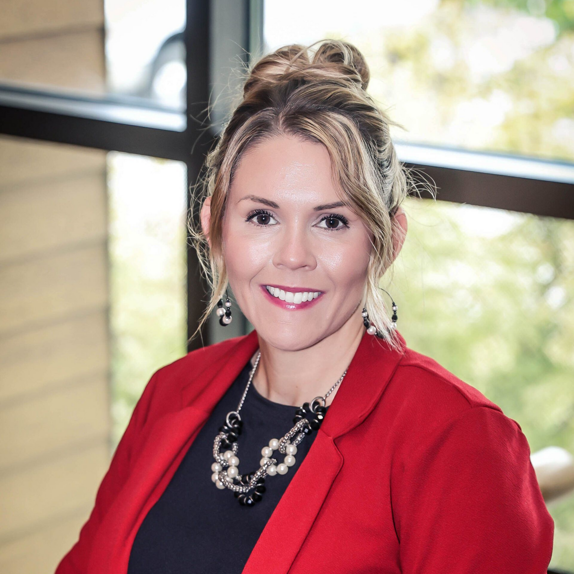 Woman in red blazer smiles. Black top, pearl necklace, hair up. Indoor setting with window.