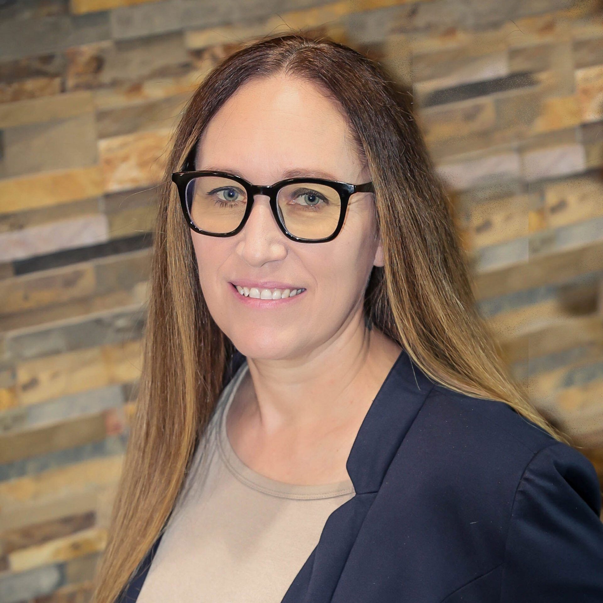 Woman wearing glasses and a blazer smiles in front of a textured wall.