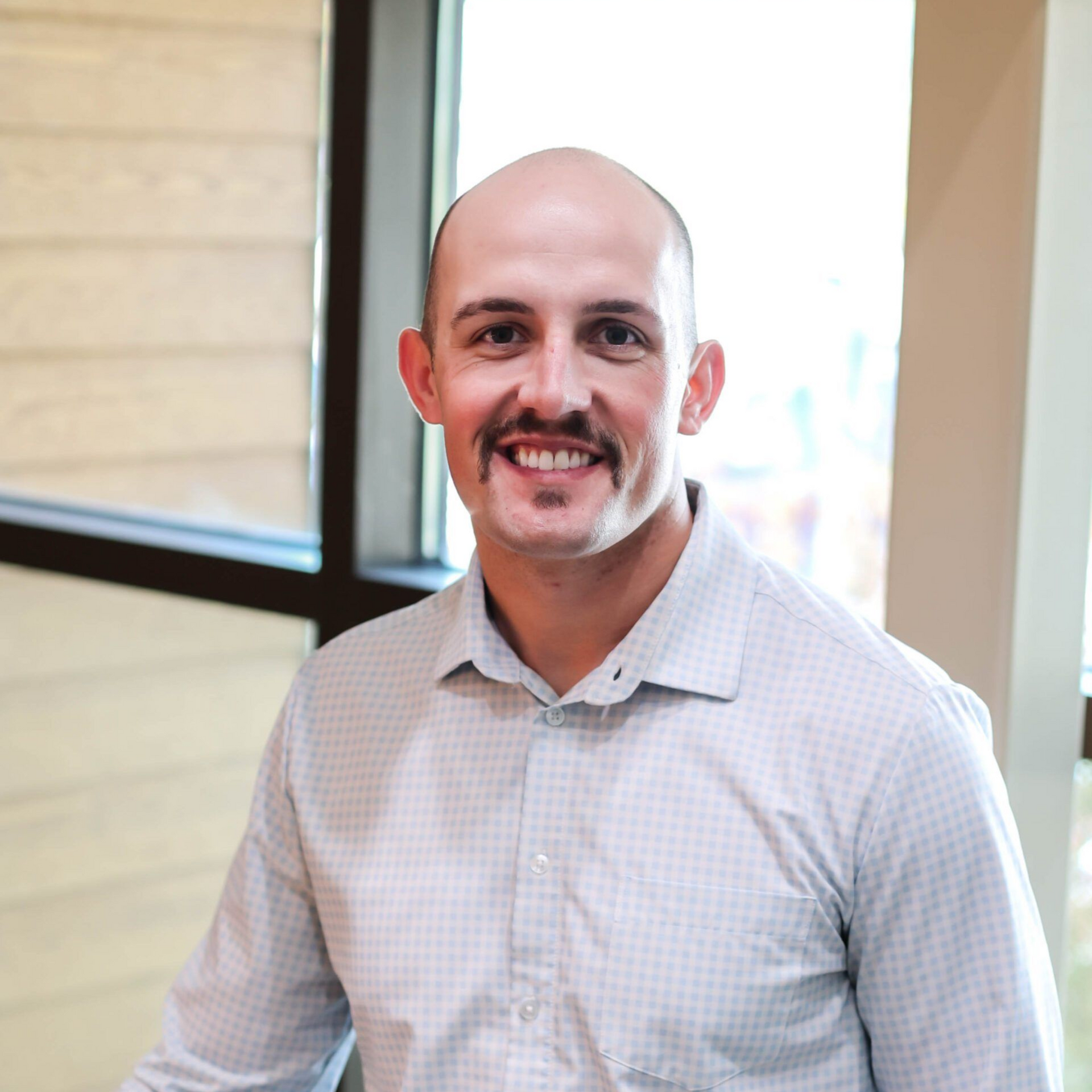 Man with mustache smiling, in a light-colored button-up shirt, standing near a window.