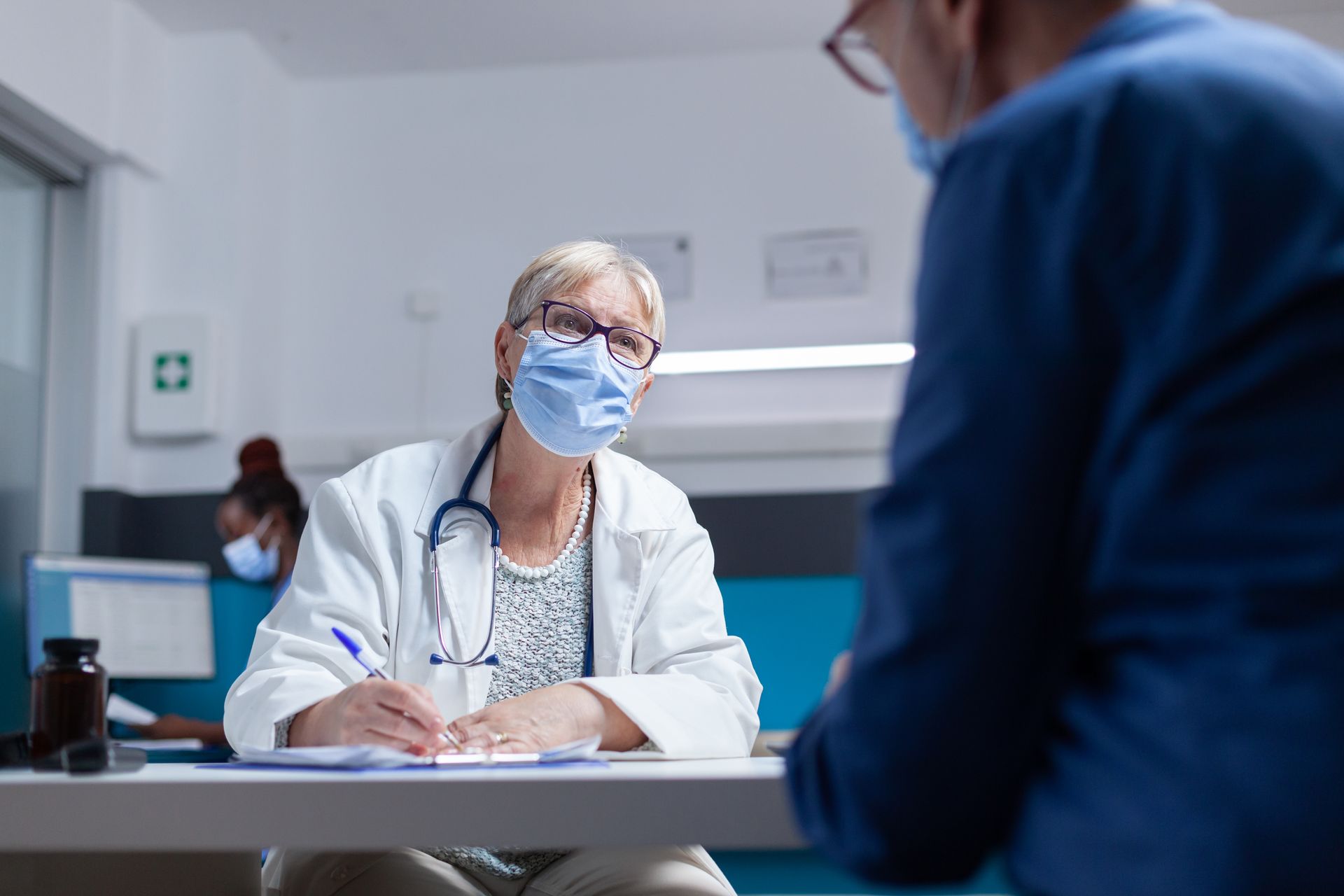 Masked doctor taking notes while speaking with a patient in a clinic office