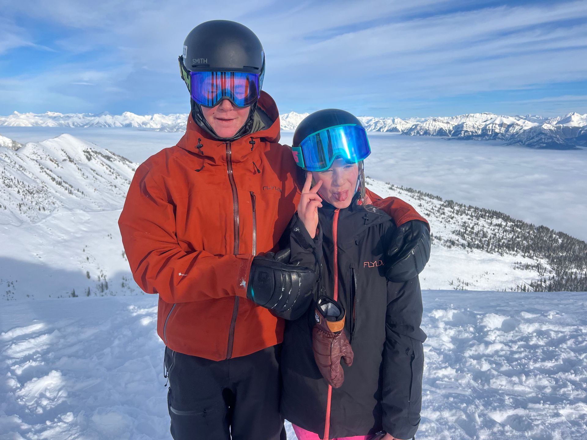 Two people in ski gear on a snowy mountaintop. Man in orange jacket and child in black jacket pose, smiling.