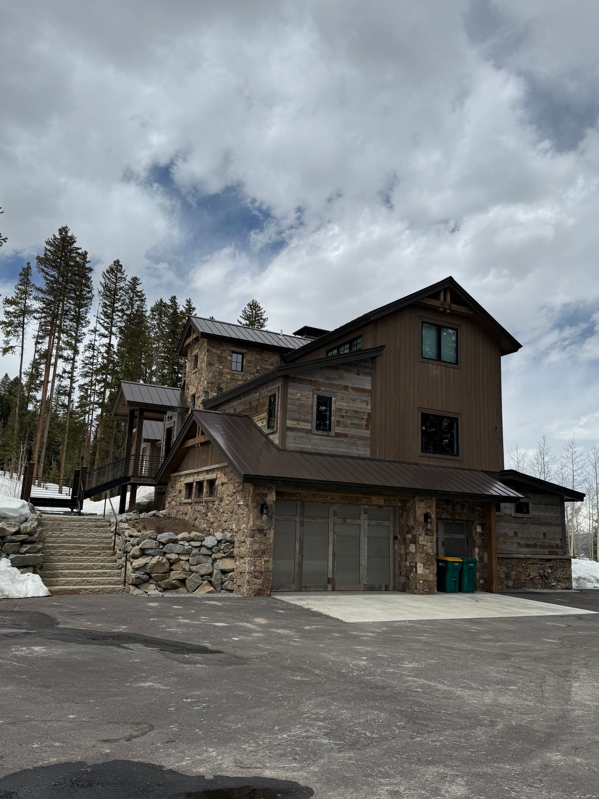 Multi-story cabin with stone and wood exterior, two-car garage, and snow-covered ground under a cloudy sky.