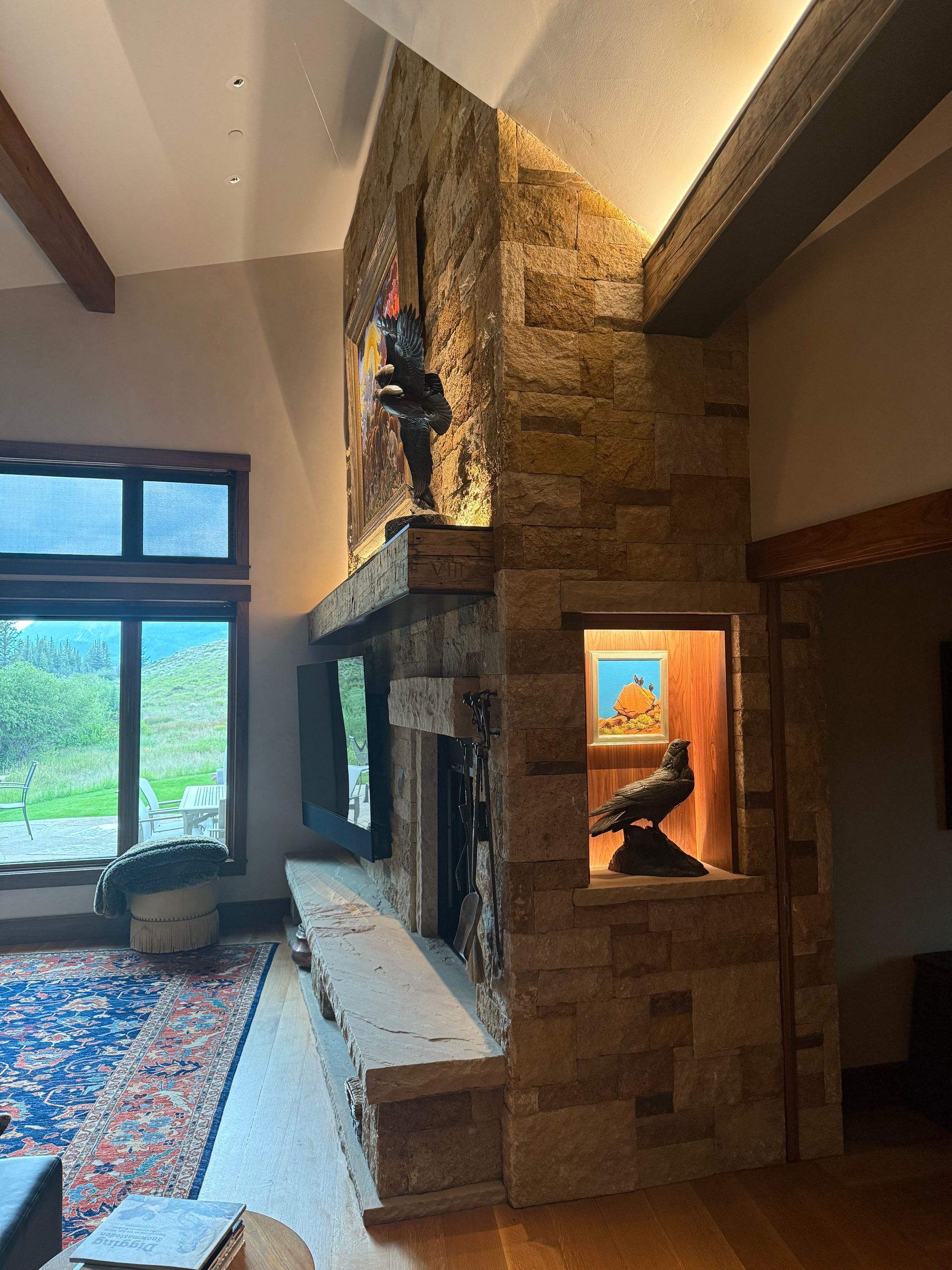 Living room with stone fireplace, built-in display of birds, large windows, and area rug.