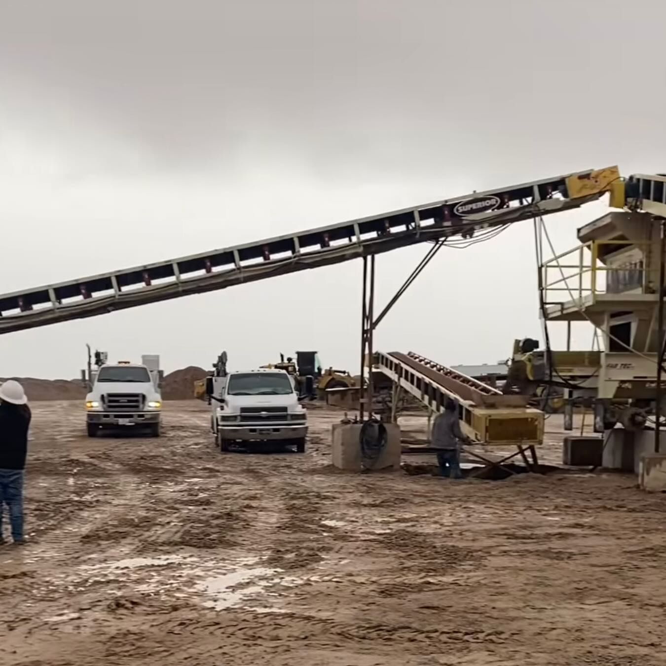 Construction site with conveyor belts and trucks under an overcast sky.