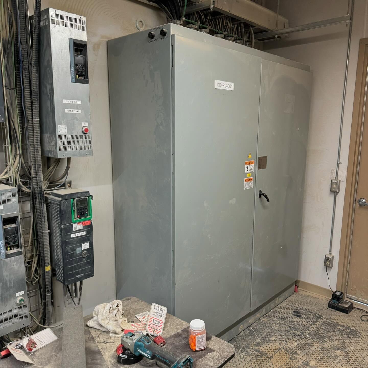 Large gray electrical cabinet in a room, with control panels and equipment on the wall. Tools are on the floor.